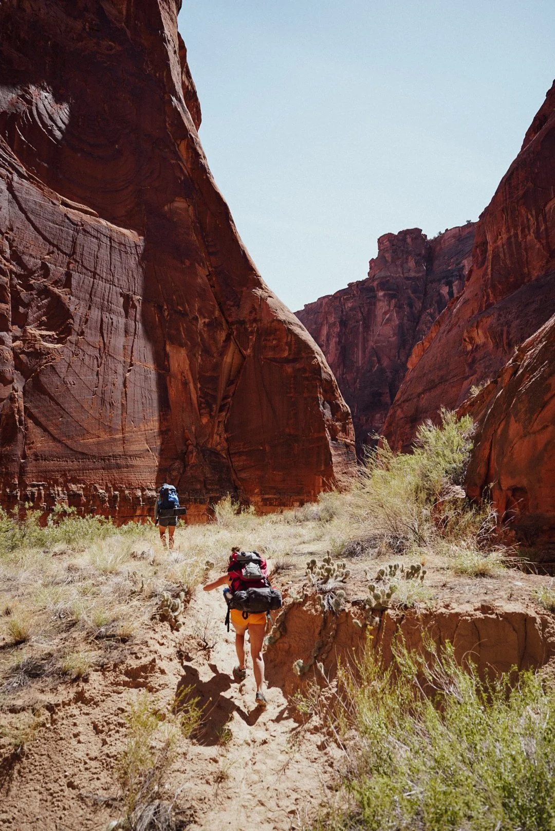 Two hikers with large backpacks walking along a dirt trail through a narrow canyon with tall red rock walls on either side.