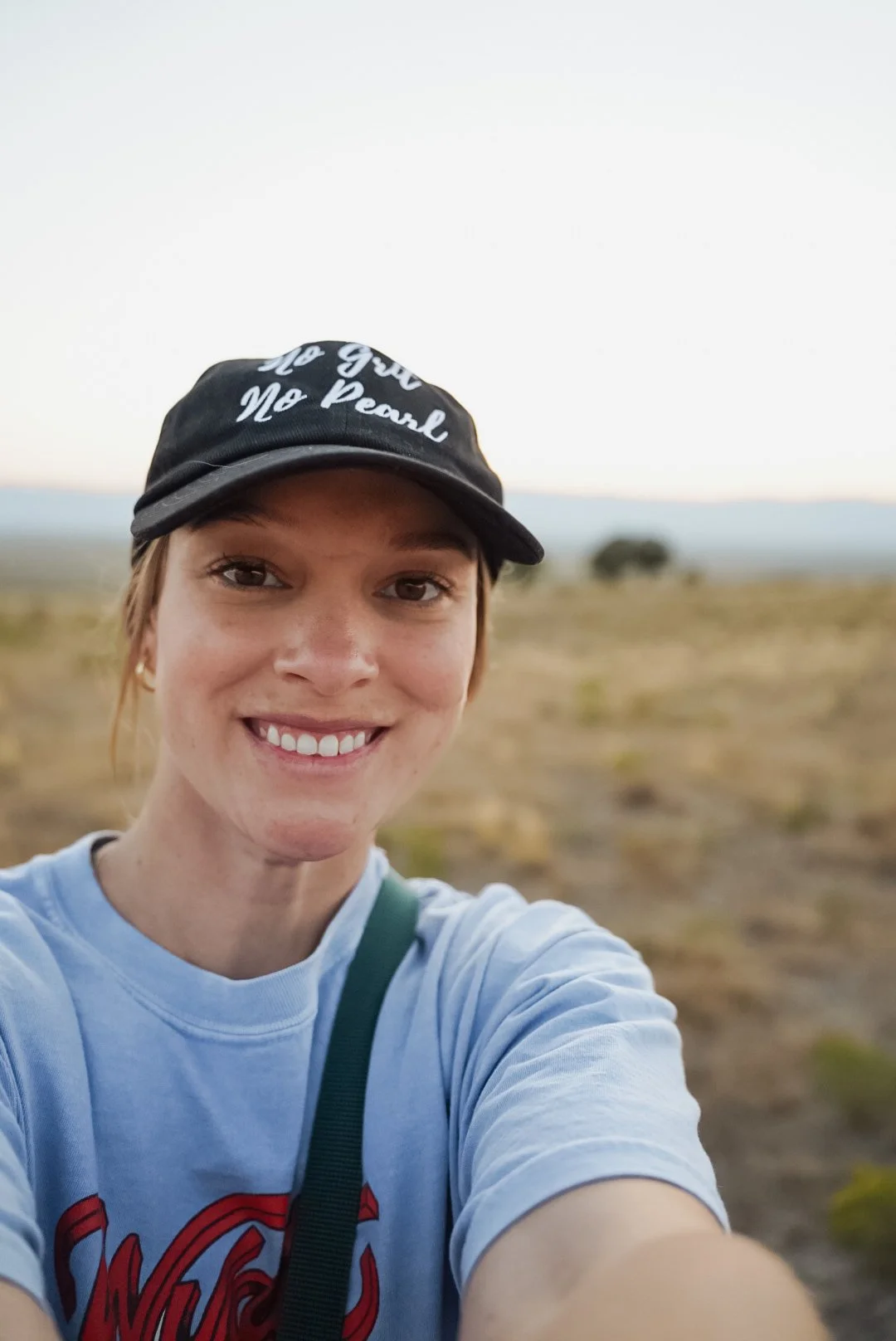 A young woman taking a selfie outdoors in a natural landscape, wearing a black cap and a light-colored T-shirt with a red graphic, smiling at the camera.