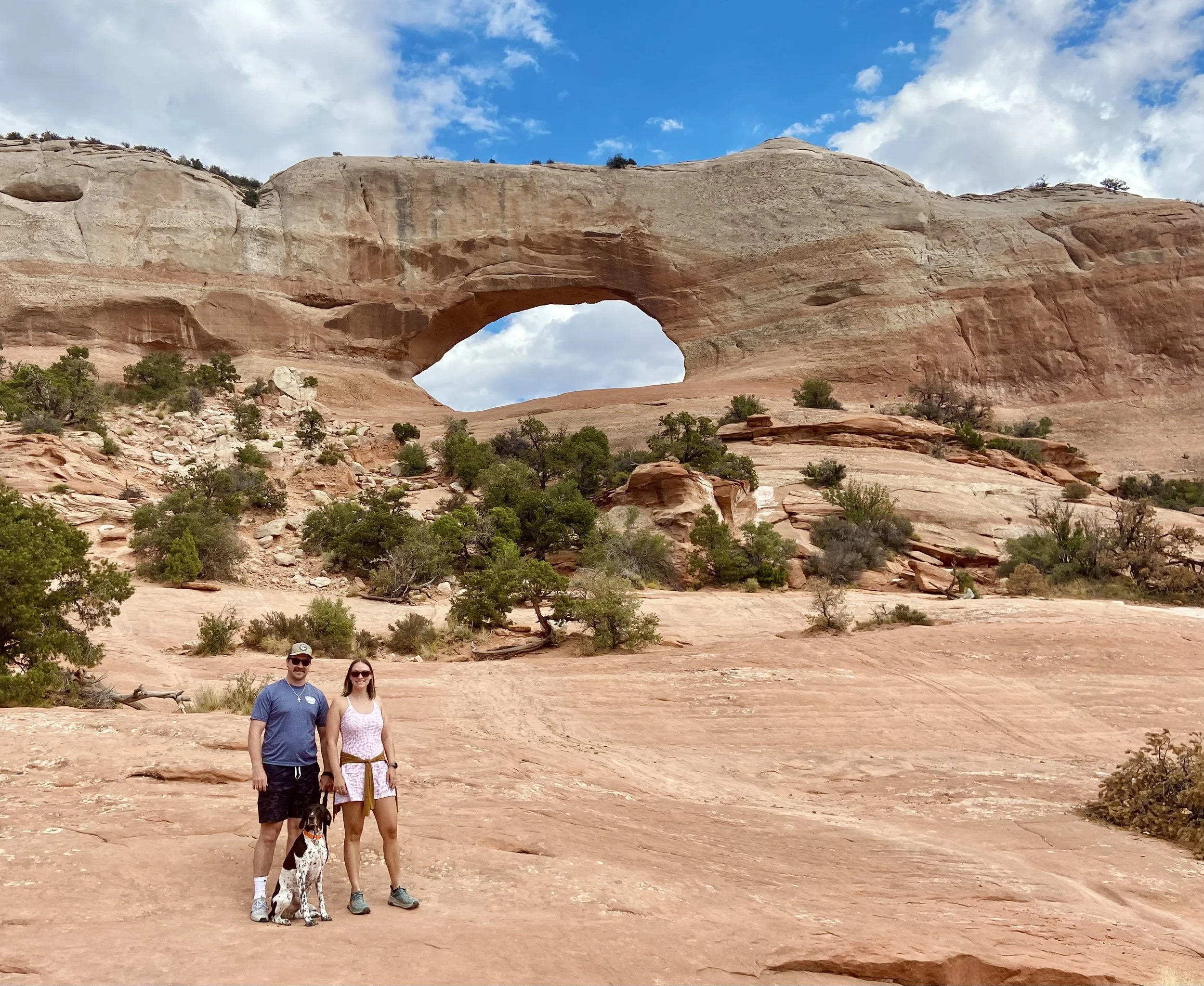 Woman, man, and dog standing in front of Wilson's Arch in Utah.