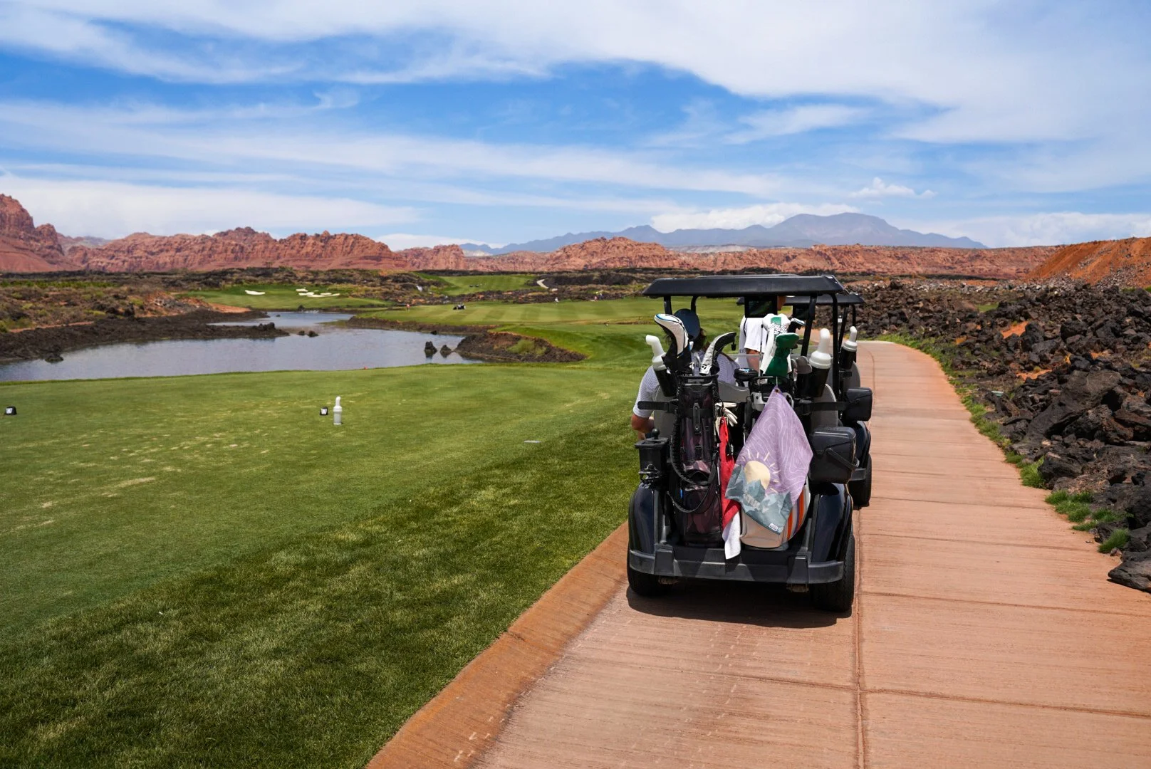 Golf cart with golf clubs on a cart passing along a paved path on a golf course with mountains and a lake in the background.