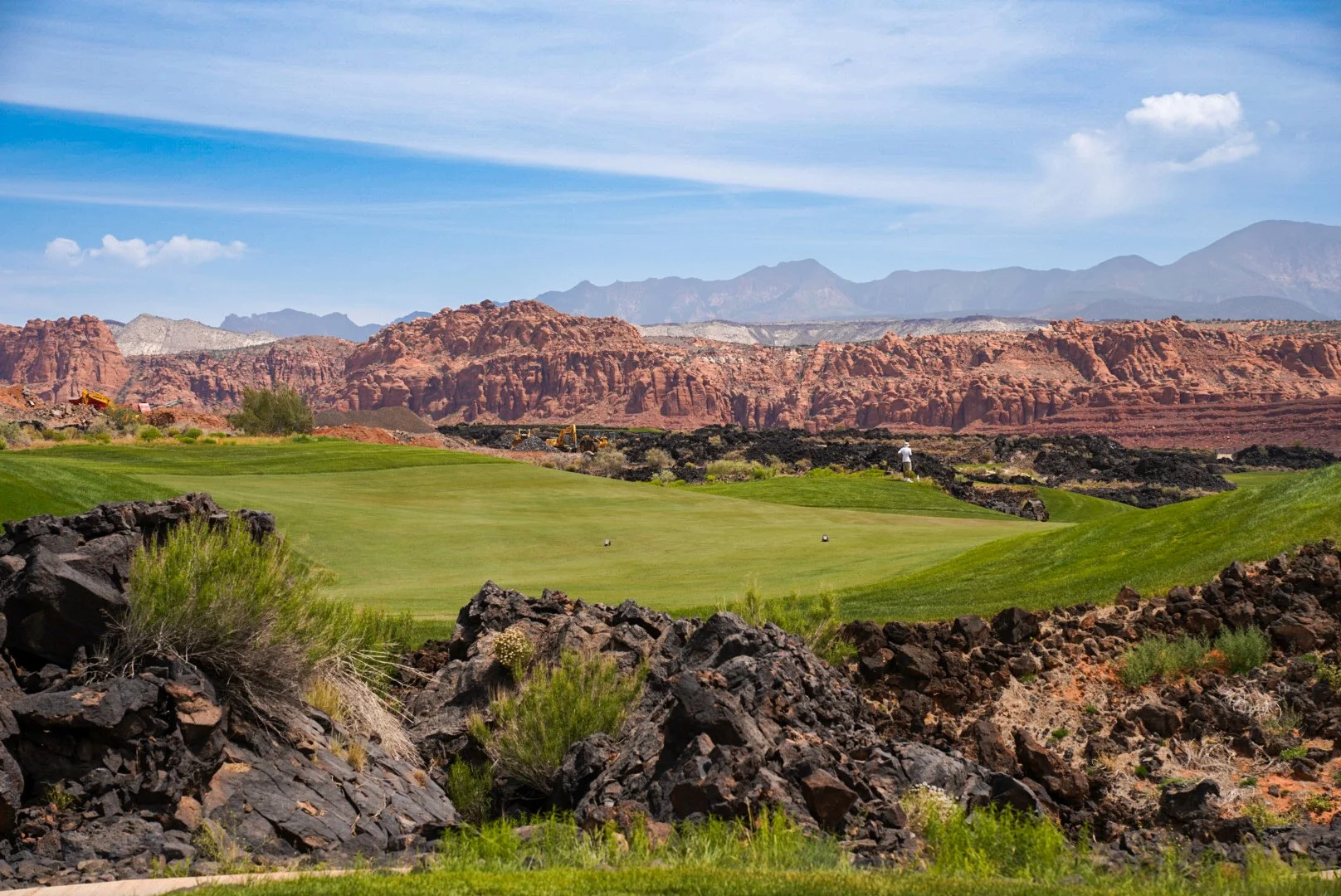 A scenic golf course with lush green grass and a lone golfer, set against a backdrop of red rock formations, mountains, and a partly cloudy sky.