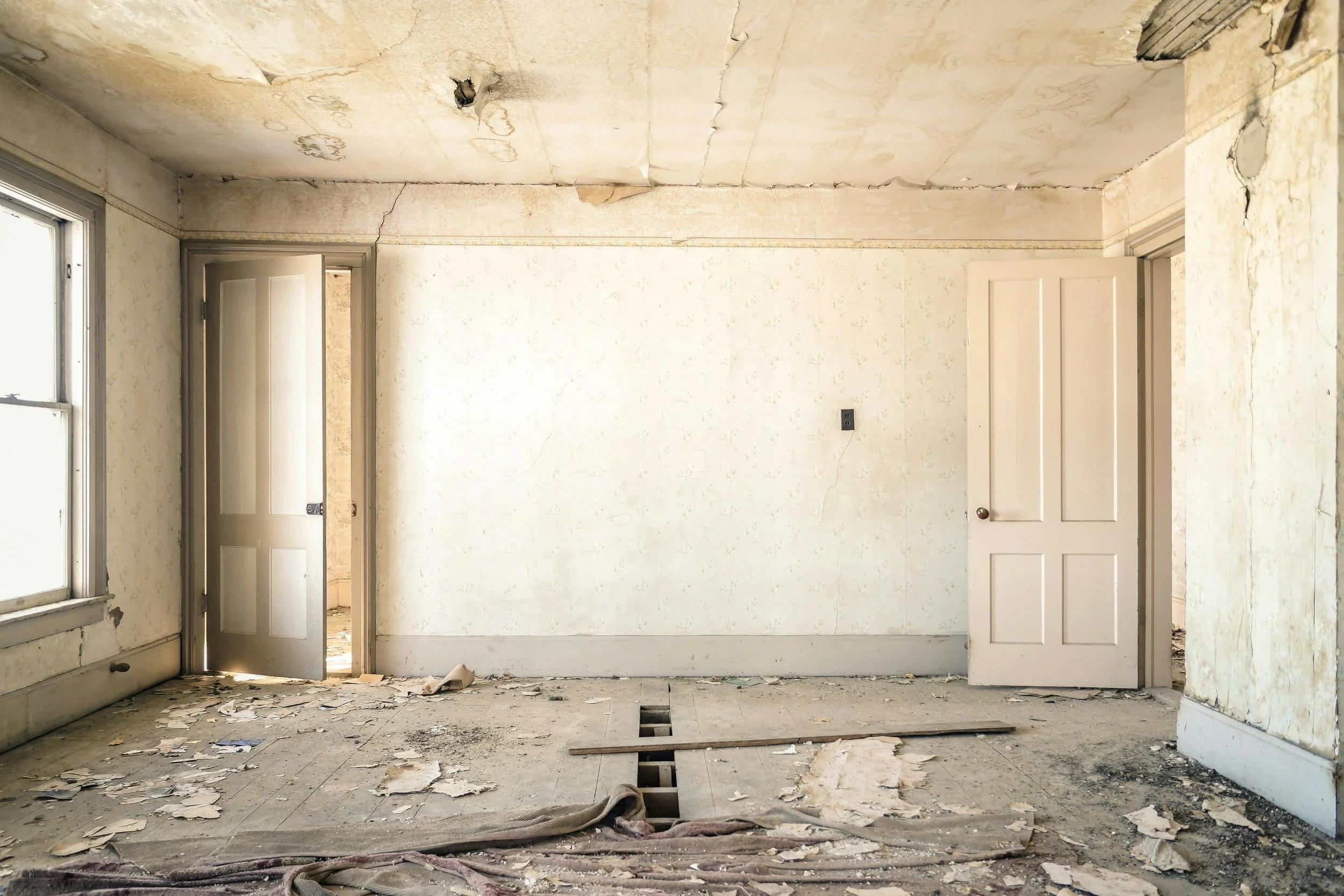 An interior room in a state of disrepair with peeling paint, a damaged ceiling, and debris on the floor. There are two doorways, one on the left and one on the right, and a window on the left wall.