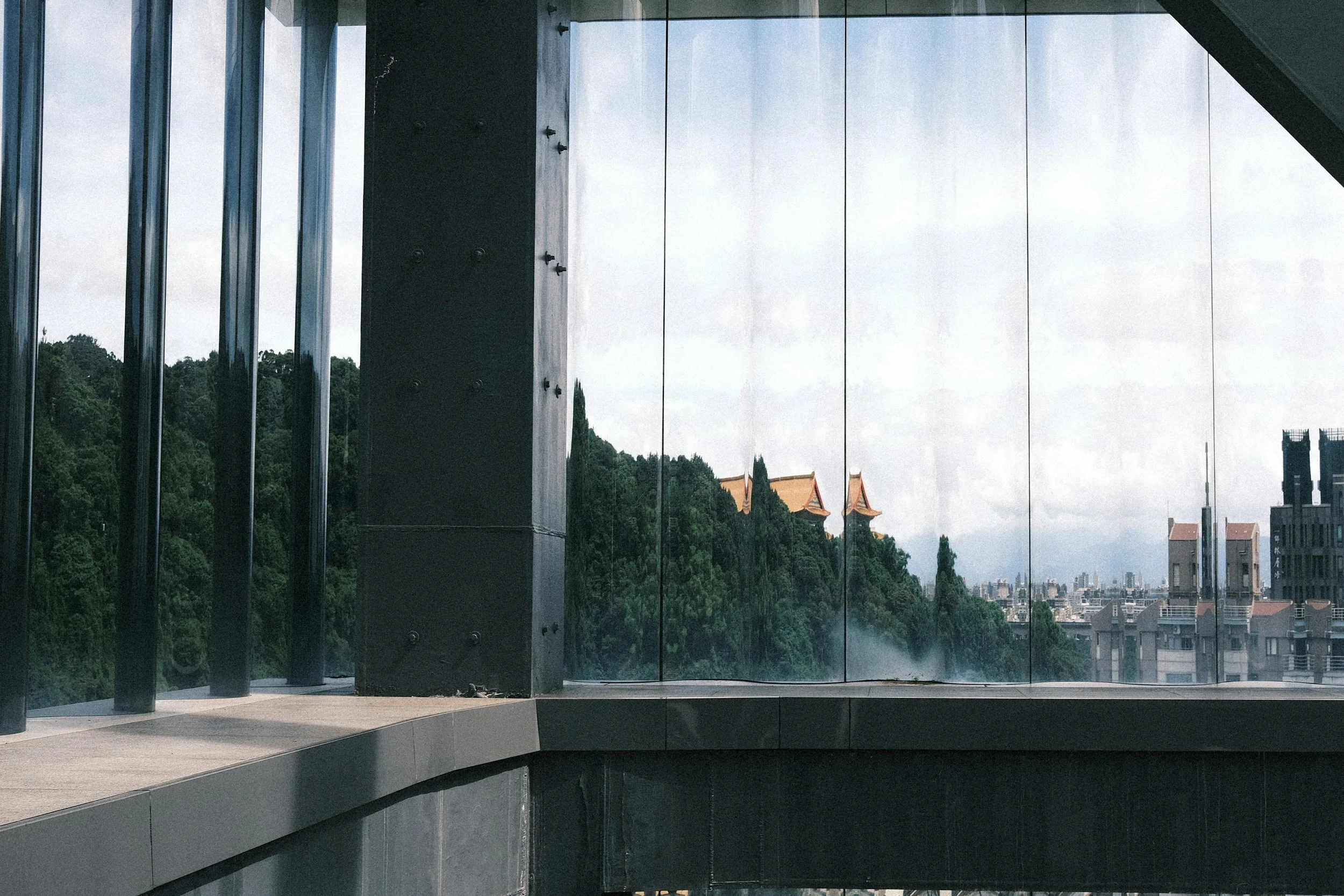 View through a large glass window showing a cityscape with a hillside covered in trees and traditional Chinese-style buildings with orange roofs, and a modern city skyline in the distance.