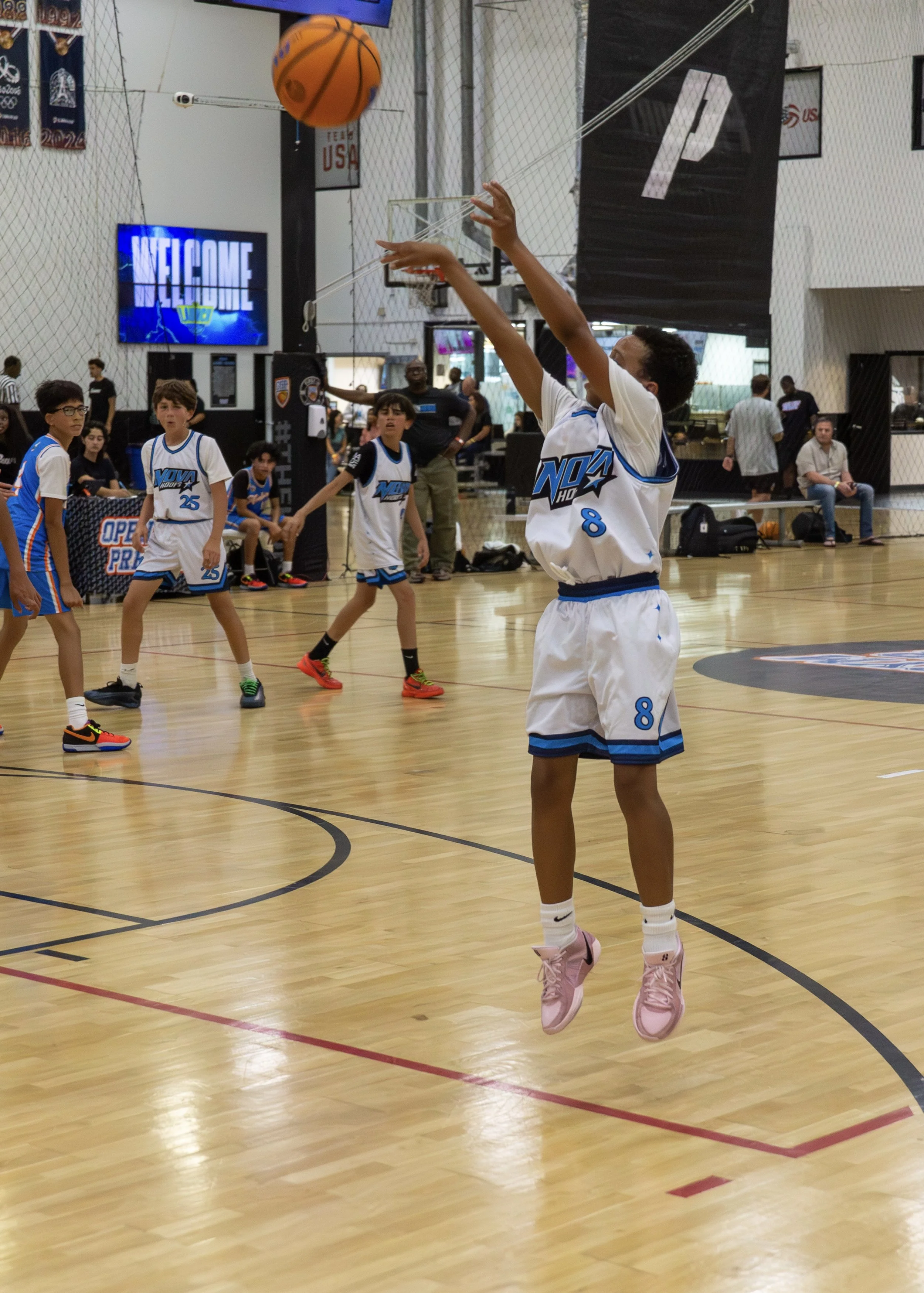 A young Nova Hoops AAU basketball player in a white jersey with blue accents and pink sneakers elevates for a jump shot during an indoor game, while teammates and other youth athletes compete and watch in the background in the Temecula–Murrieta area.