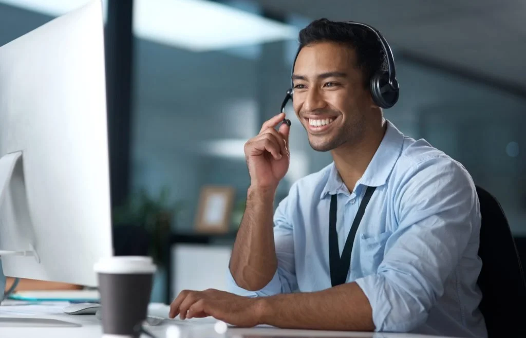 A man with a headset smiling at a computer in an office environment.