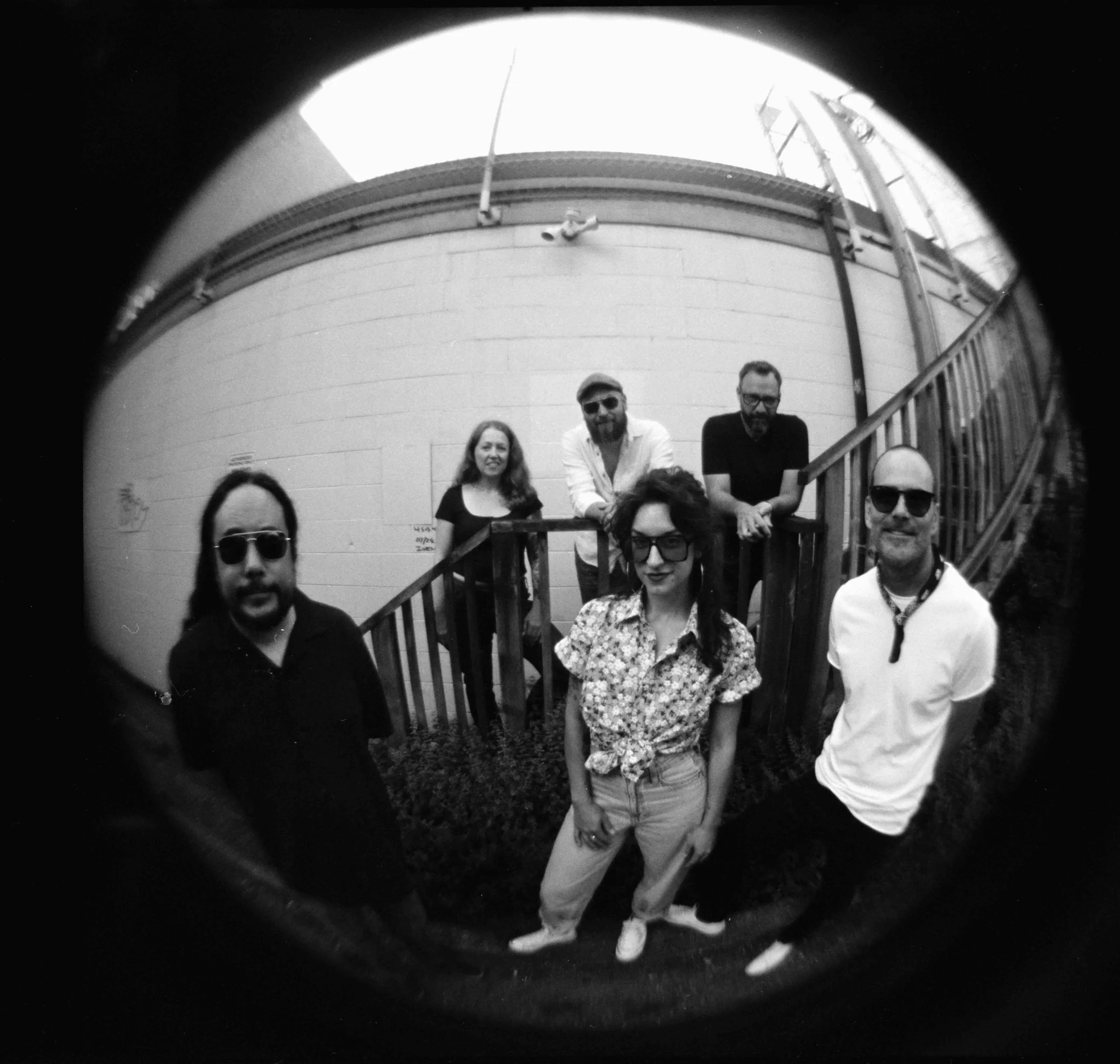 Black and white photo of six people posed outside, with a wooden staircase and railing behind them. The group consists of three men and three women, dressed casually, some wearing sunglasses, standing in front of a brick building.