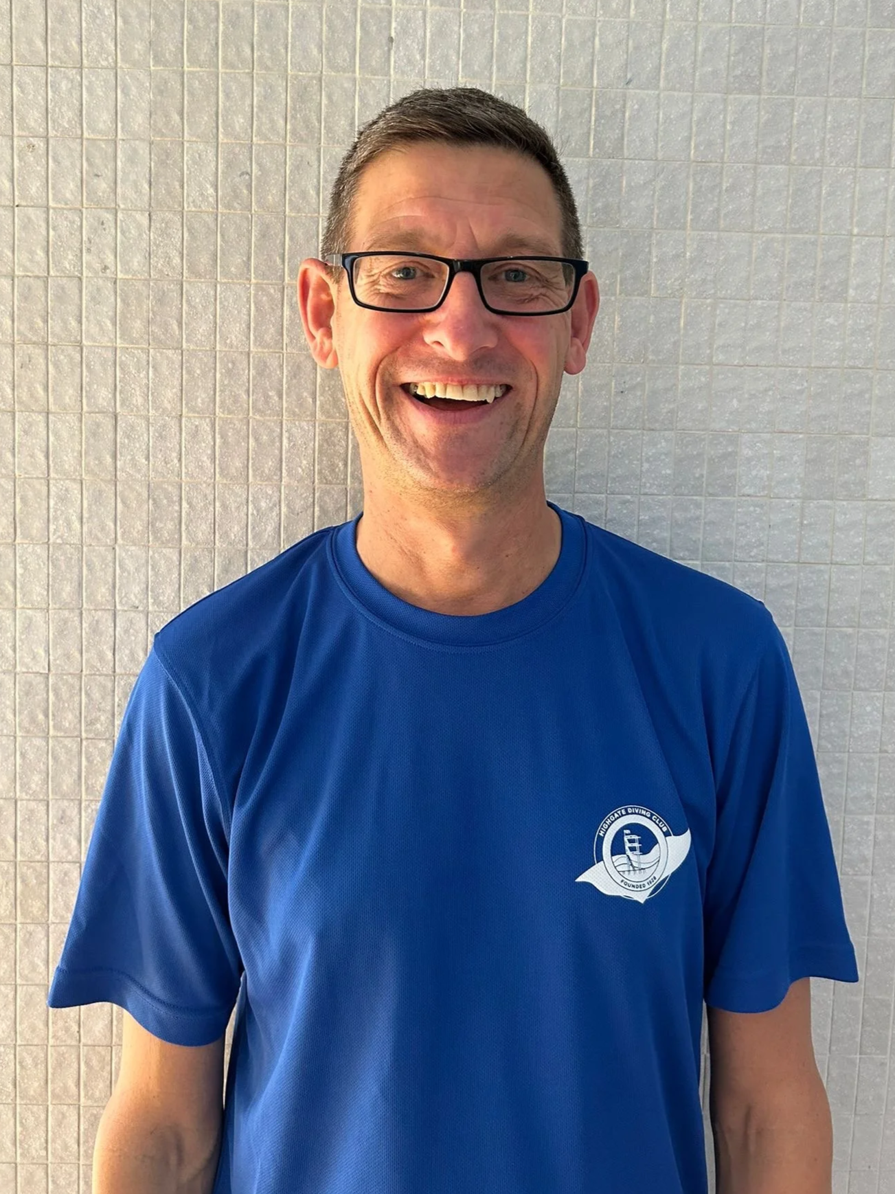 A happy man with glasses smiling in front of a tiled wall, wearing a blue sports shirt with a logo on the chest.