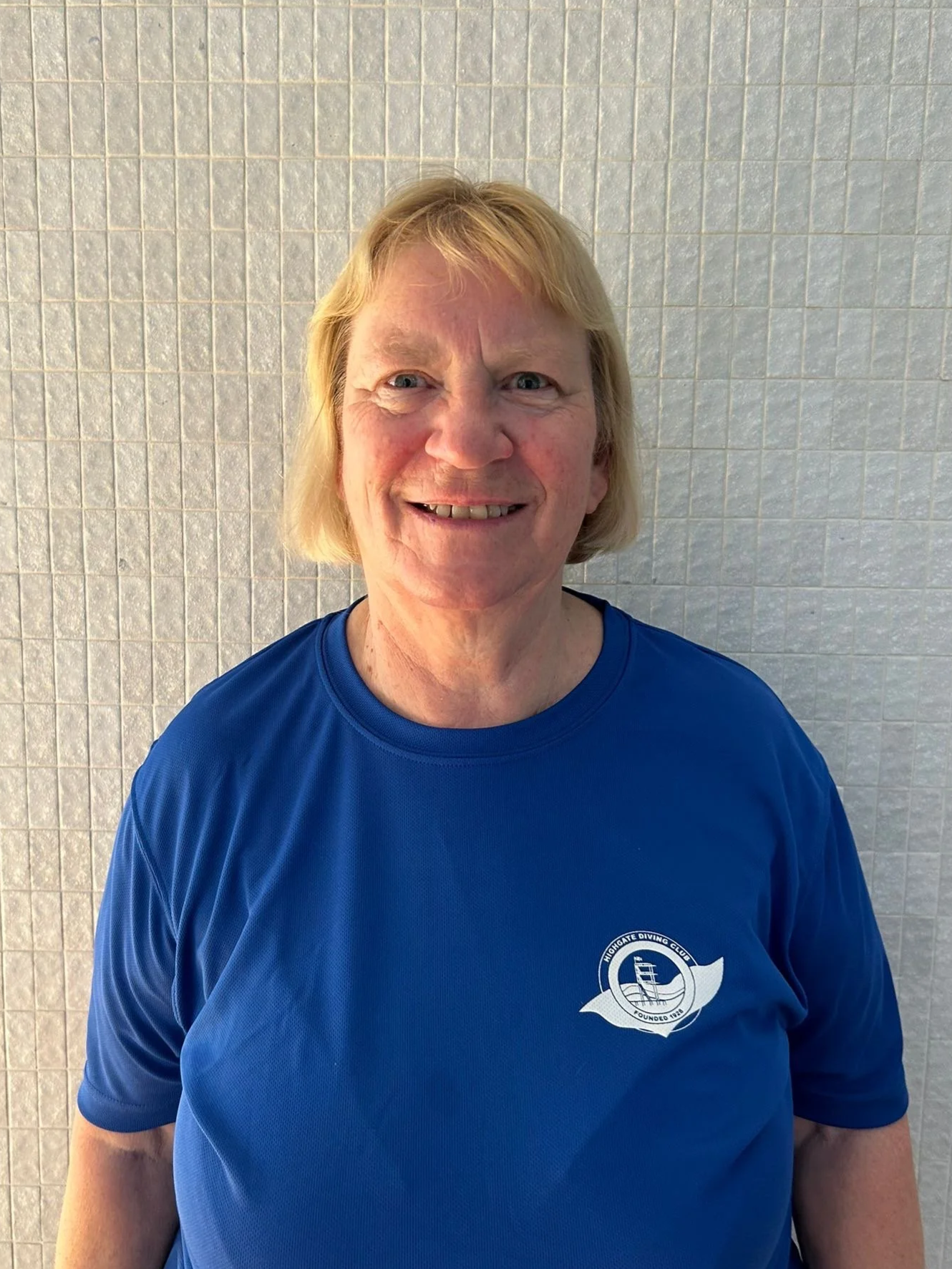A smiling woman with blonde hair wearing a blue T-shirt with a logo, standing in front of a tiled wall.