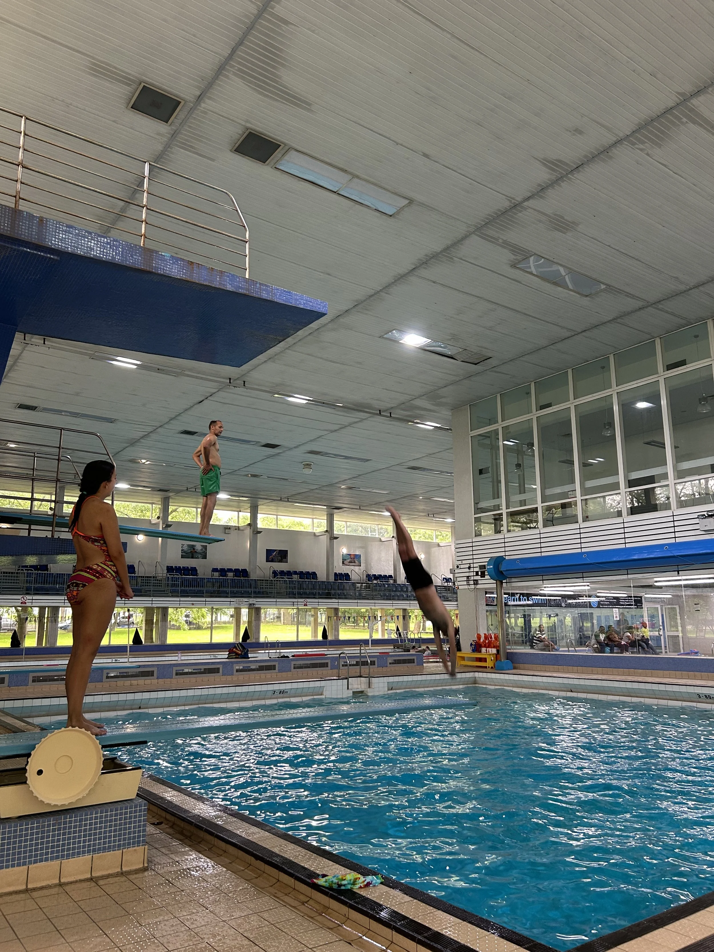 An indoor swimming pool with a diver mid-air diving into the water, a person standing on the diving board, and another person observing. The pool area has large windows and seating areas for spectators.