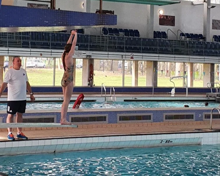 A woman performing a standing dive on the edge of an indoor swimming pool while a man watches nearby.