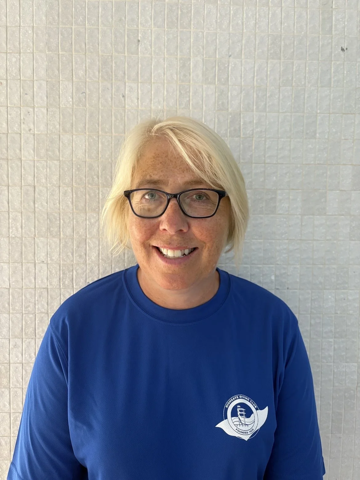 Close-up of a young woman with short, red hair and blue eyes, standing outdoors near a body of water with mountains in the background.