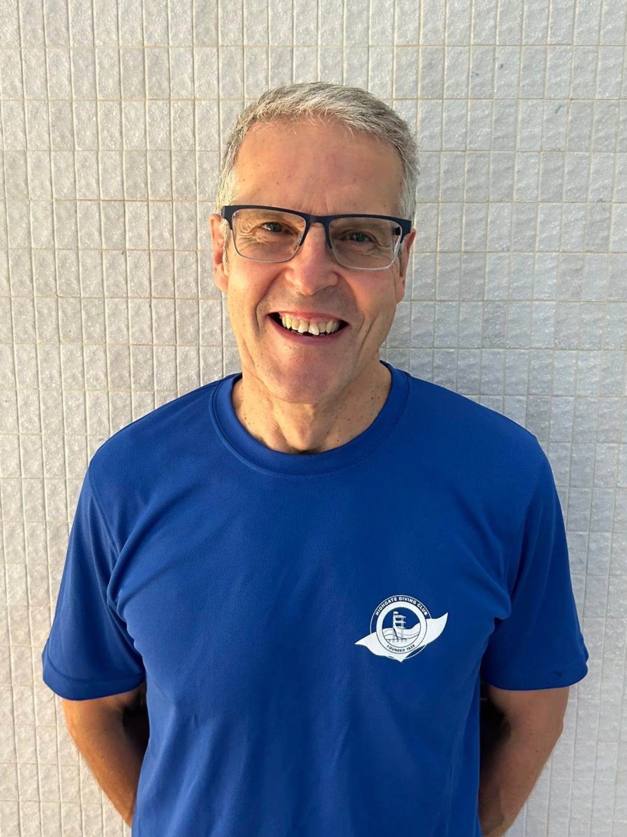 Smiling man with glasses wearing a blue t-shirt with a logo of a boat on the chest, standing in front of a tiled wall.