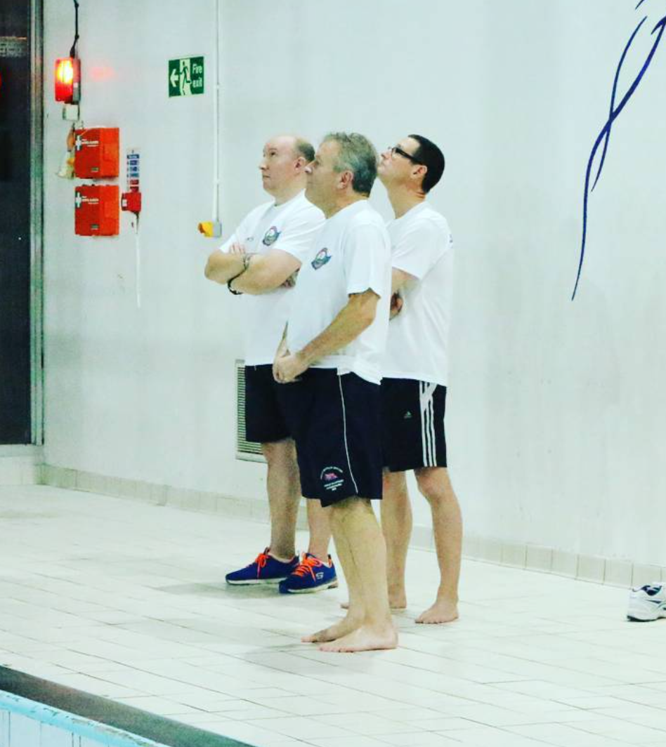 Three men standing in line at the edge of a swimming pool, wearing white T-shirts and black shorts, looking towards an emergency light and fire equipment on the wall.
