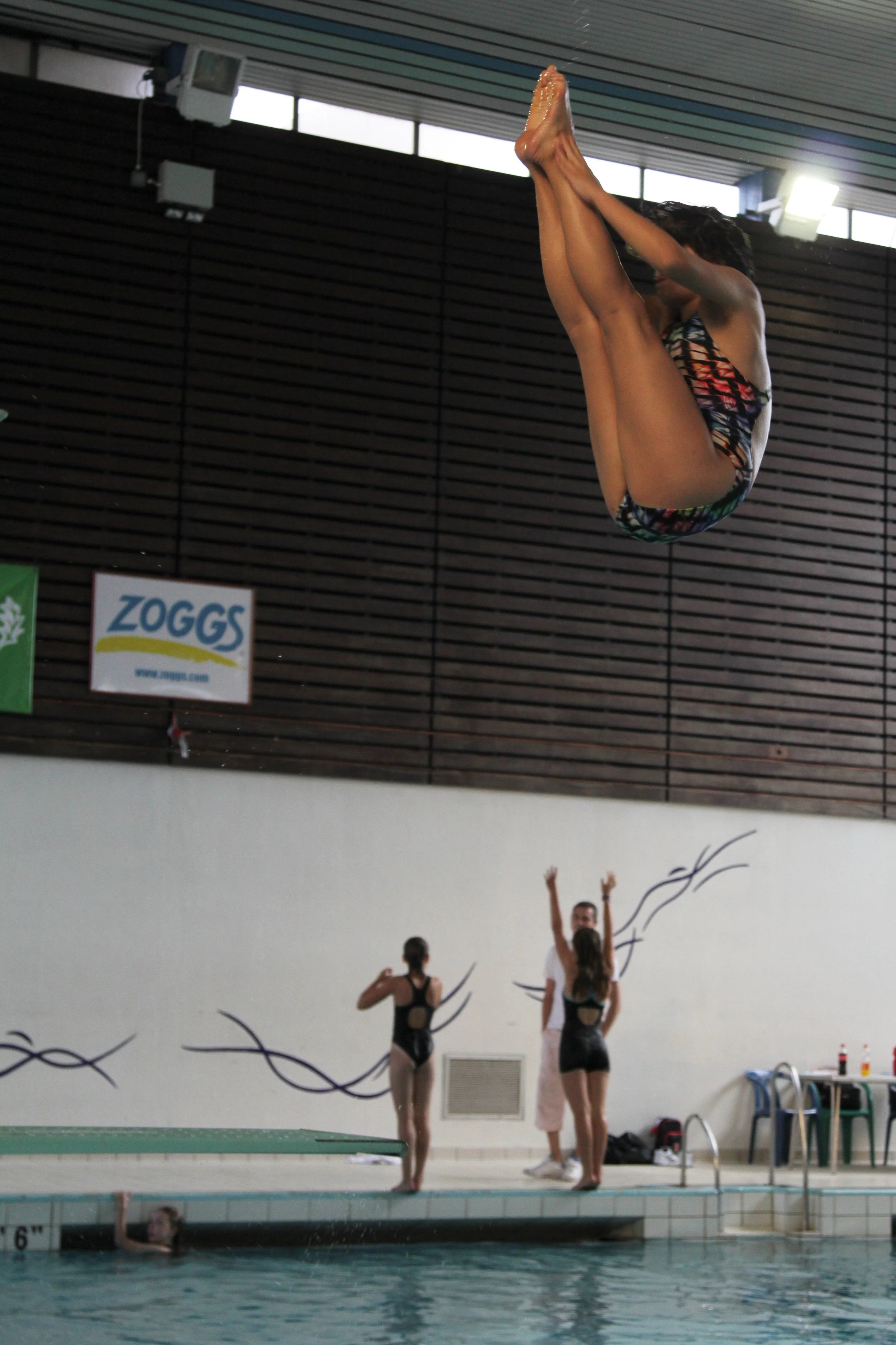 A young girl performing a backwards dive pike into a swimming pool, with other swimmers and a coach watching in the background at White Oak Leisure Centre.