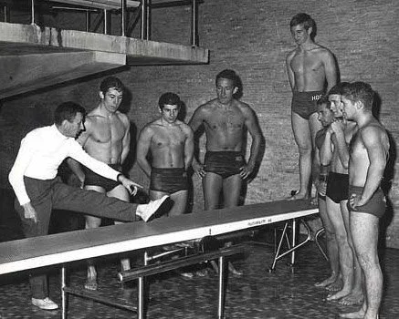 A male coach demonstrating stretching techniques to a group of six young male athletes in swimwear, standing around a poolside in an indoor swimming pool area.