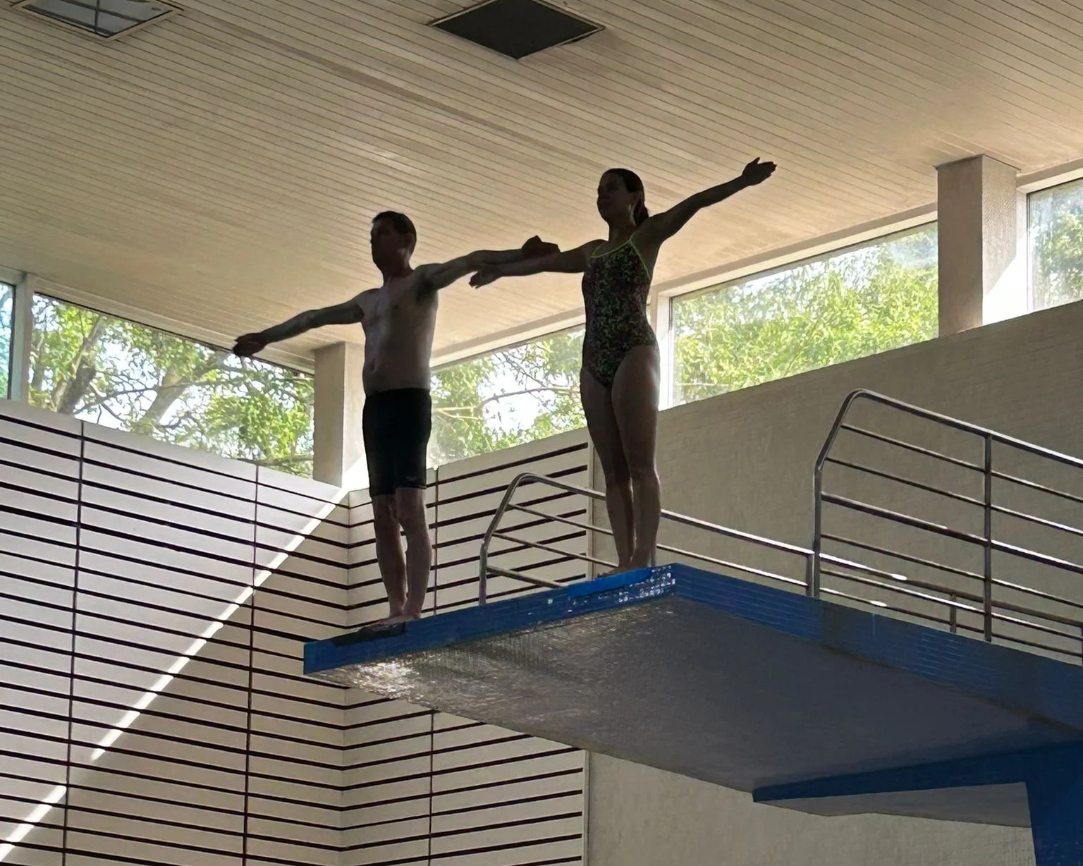Two synchronized divers practicing on a springboard in an indoor pool area with large windows showing trees outside.