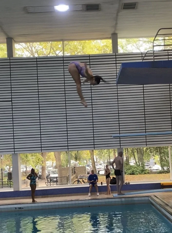 A female diver in a purple swimsuit is mid-air above a swimming pool, diving from a platform into the water, with several people observing nearby.