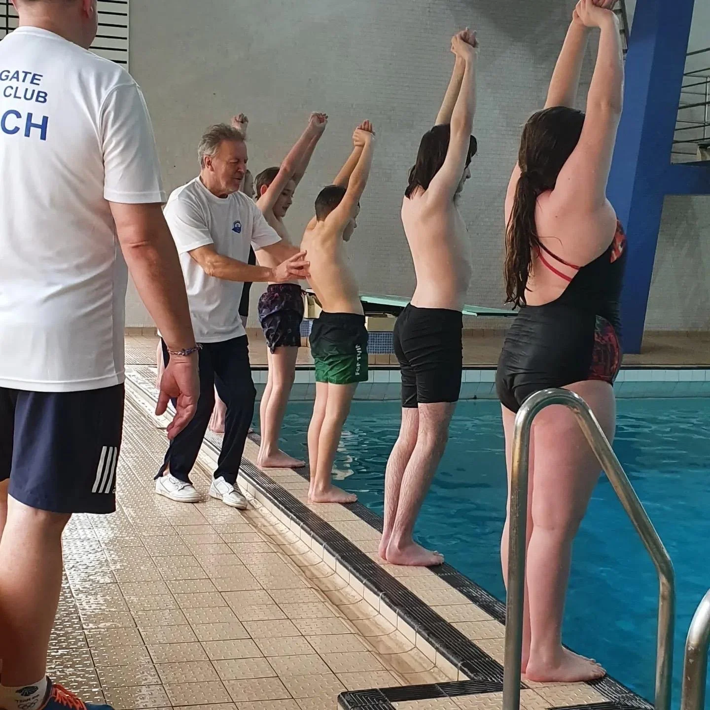 People standing on the edge of an indoor swimming pool, preparing to jump or dive into the water, with an instructor guiding them.