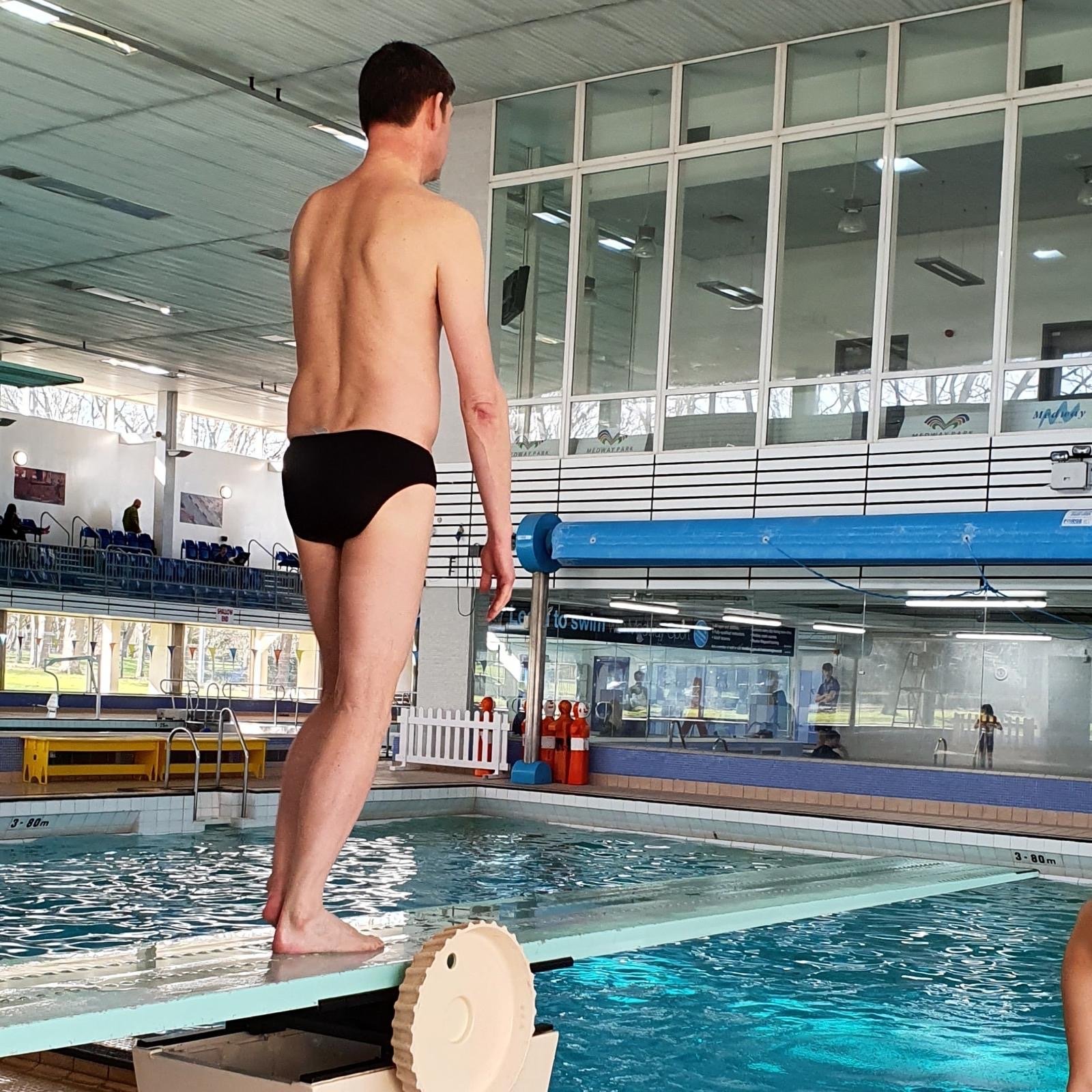 A man in black swim briefs standing on the diving board at an indoor swimming pool, preparing to jump.
