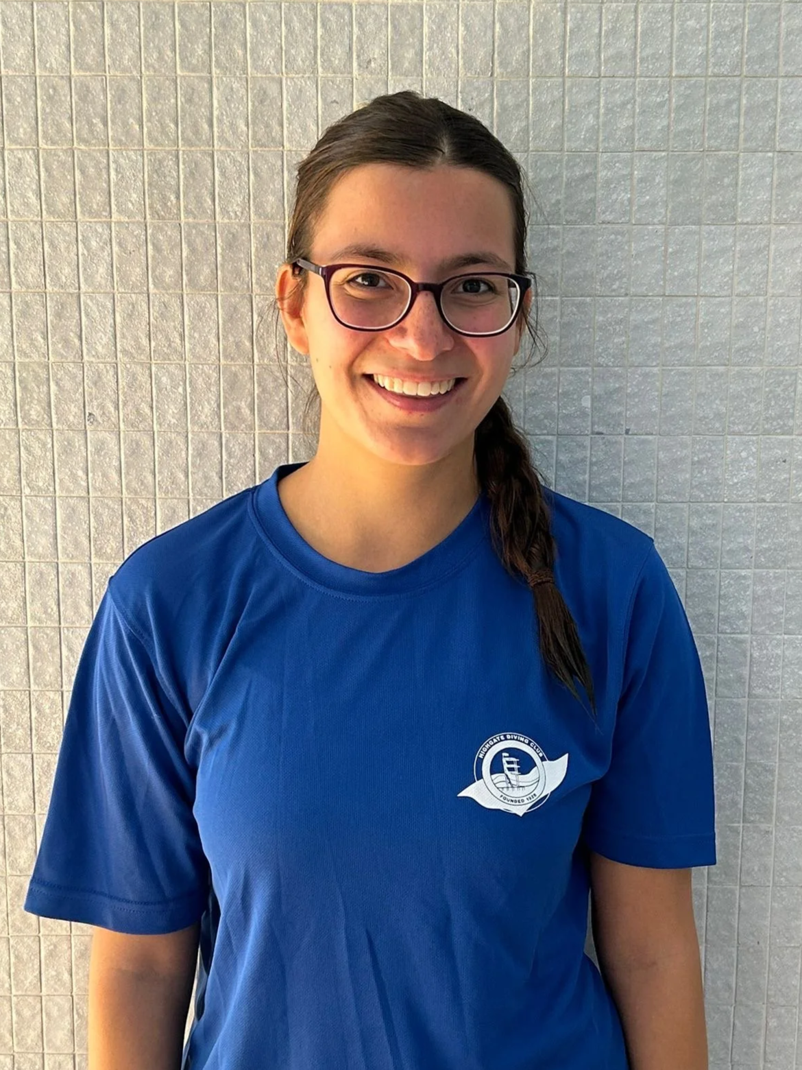 Smiling young woman with glasses and a braided hair, wearing a blue T-shirt with a logo, standing against a tiled wall.