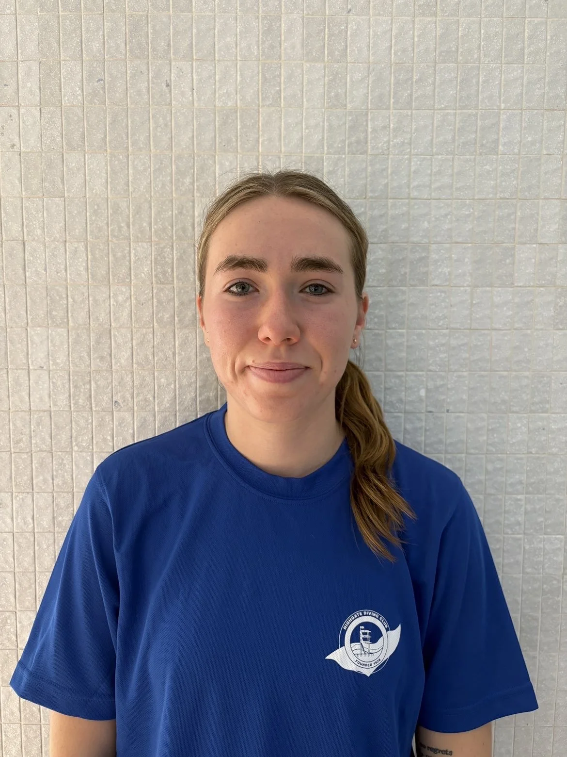 A young woman with blonde hair and blue eyes looking directly at the camera. She is sitting outside near a rocky background with sunlight filtering through leaves, casting shadow patterns on her face and chest. She is wearing a white top with puffed sleeves.