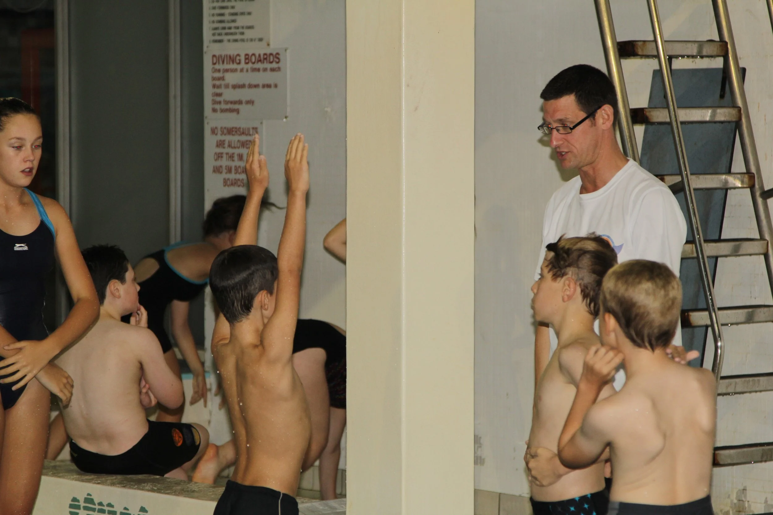 Divers listening to a coach during a diving practice at an indoor pool, with some kids raising their hands and others standing or sitting on the pool edge.