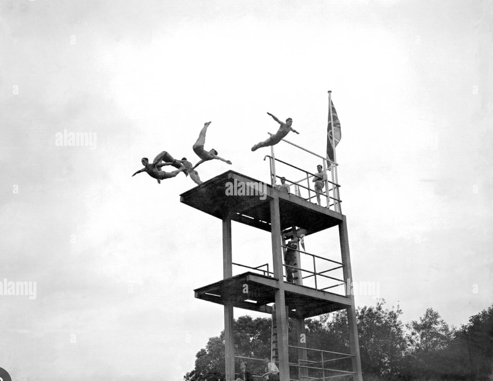 Four people performing a synchronized dive from a diving platform with two levels, a person standing on the lower level holding a flag, and a few people in the background, under a cloudy sky.