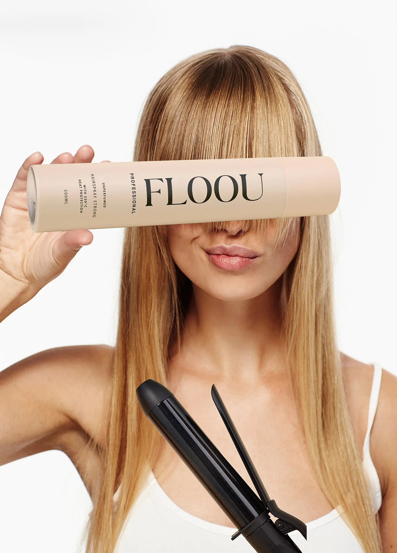 Nainen, jolla on vaaleat hiukset ja valkoinen toppi, pitää hiusten suoristusrautaa ja peittää osittain kasvonsa hiusten otsatukalla, samalla kun hän pitää hiustenhoitotuotepakettia, jossa lukee 'FLOOU'.