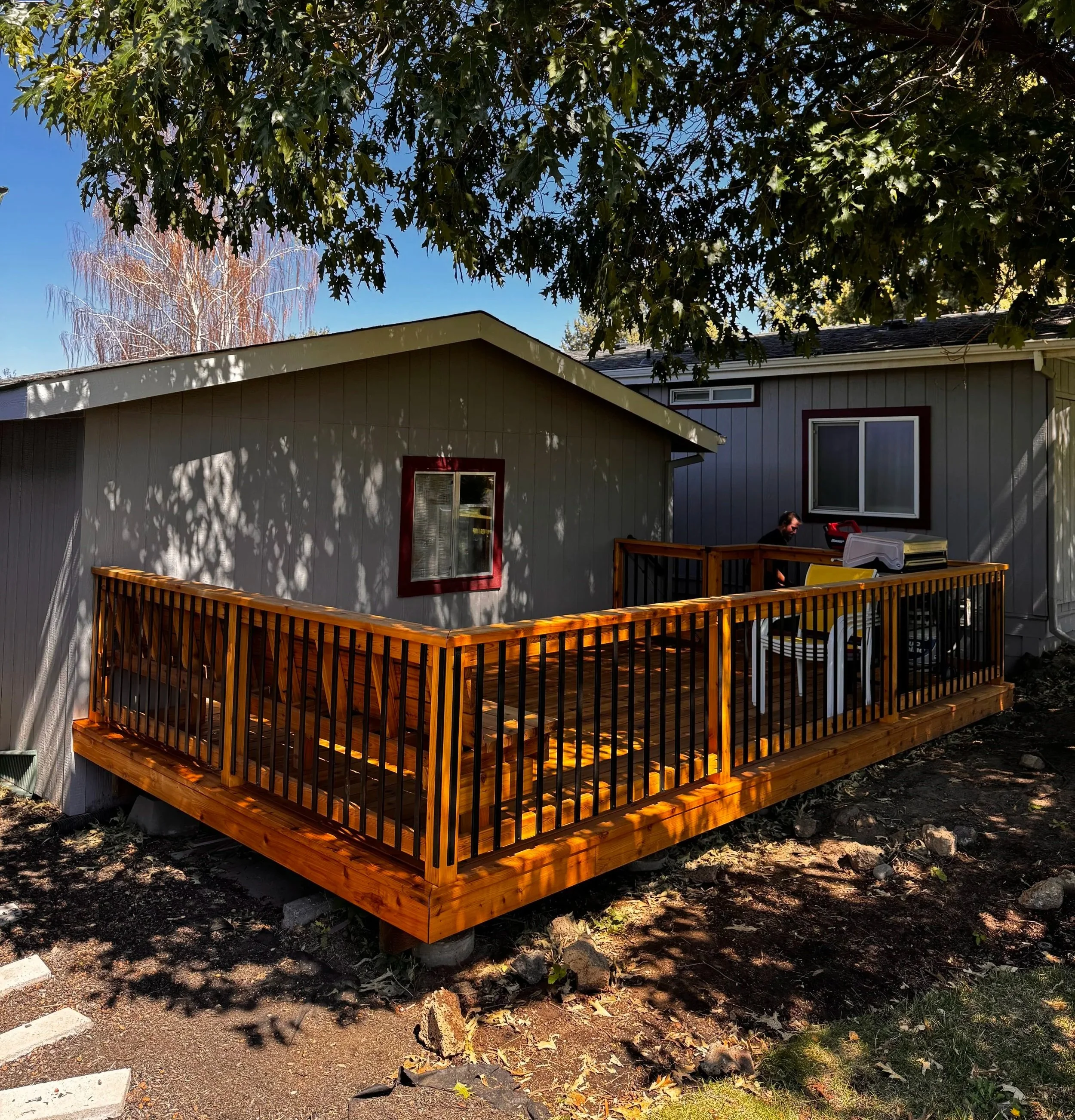 A wooden deck with railing attached to the back of a gray house with a window. A person is sitting on the deck, partially visible. There is a backyard with trees, including a large tree with green leaves overhead and a smaller tree with reddish and white branches across the yard. The ground is covered with soil and scattered leaves. A few items, including a white plastic container, are on the deck.