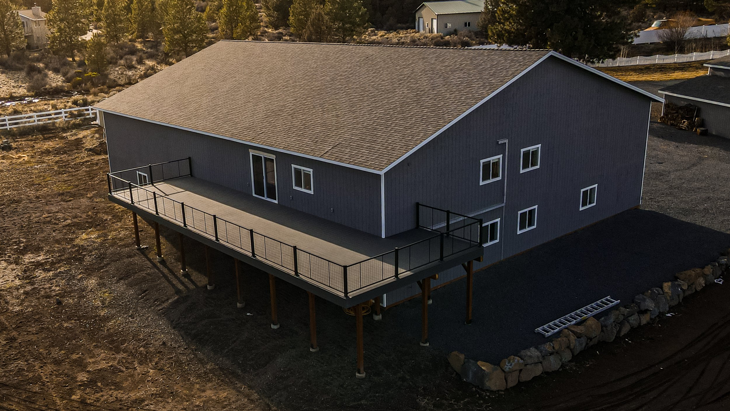 A two-story gray house with a pitched roof and multiple windows, featuring a large wooden deck with a black railing, elevated on stilts in a rural or suburban area, with other houses and trees in the background.