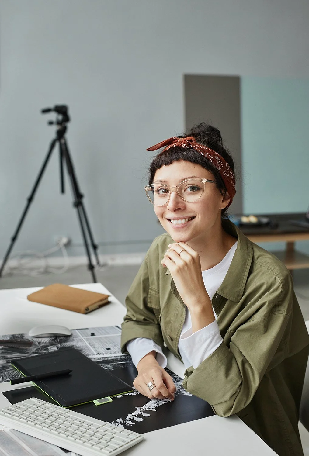 A young woman with glasses and a red bandana smiling at the camera, seated at a desk with a keyboard, a pen, notebooks, and a tablet, in a modern workspace with a tripod in the background.