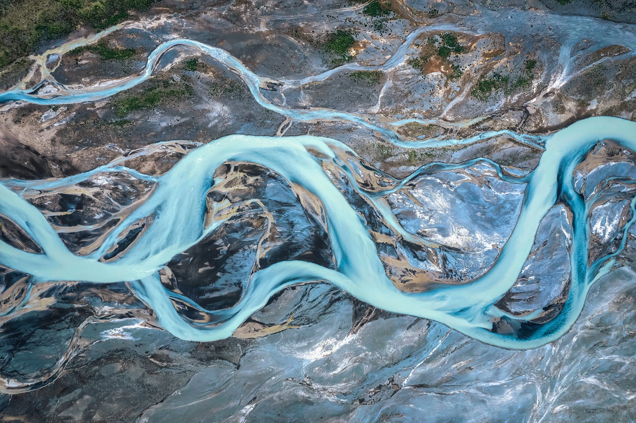 Aerial view of a winding river with light blue water flowing through a rugged landscape with patches of green vegetation and exposed earth.