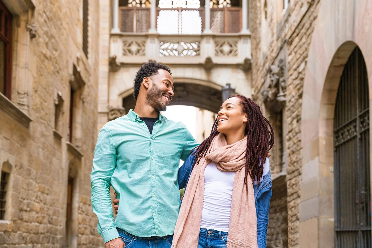 A smiling couple walking together in an alley with historic architecture, looking at each other, with the man placing his arm around the woman's shoulder.