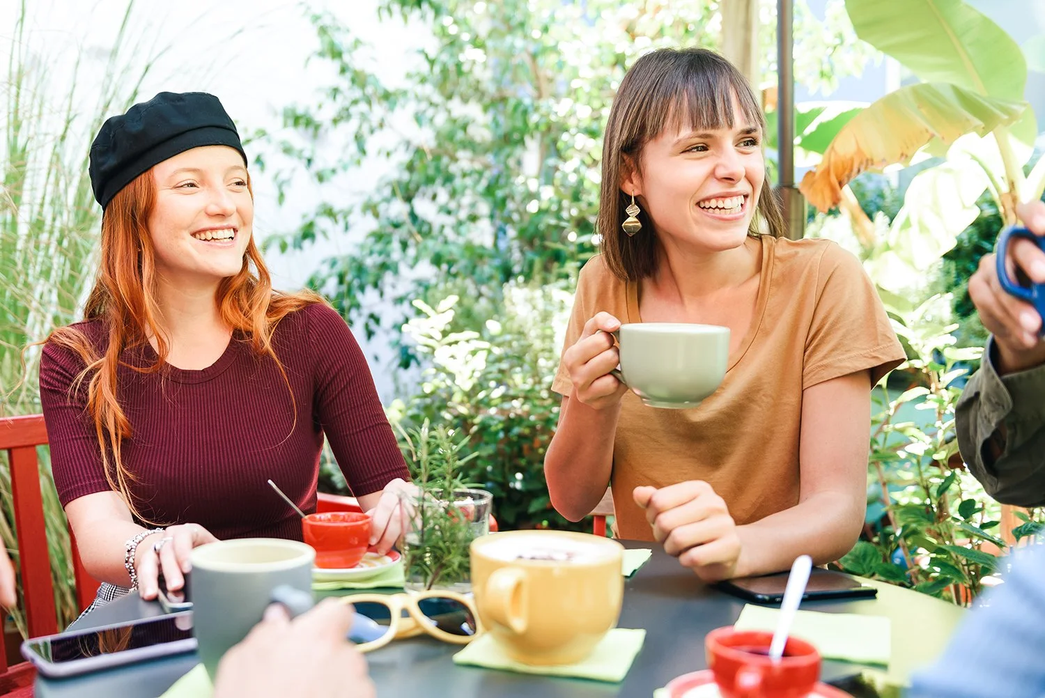 Two women sitting at a table outdoors, smiling and talking, with a woman holding a cup and a person showing scissors.