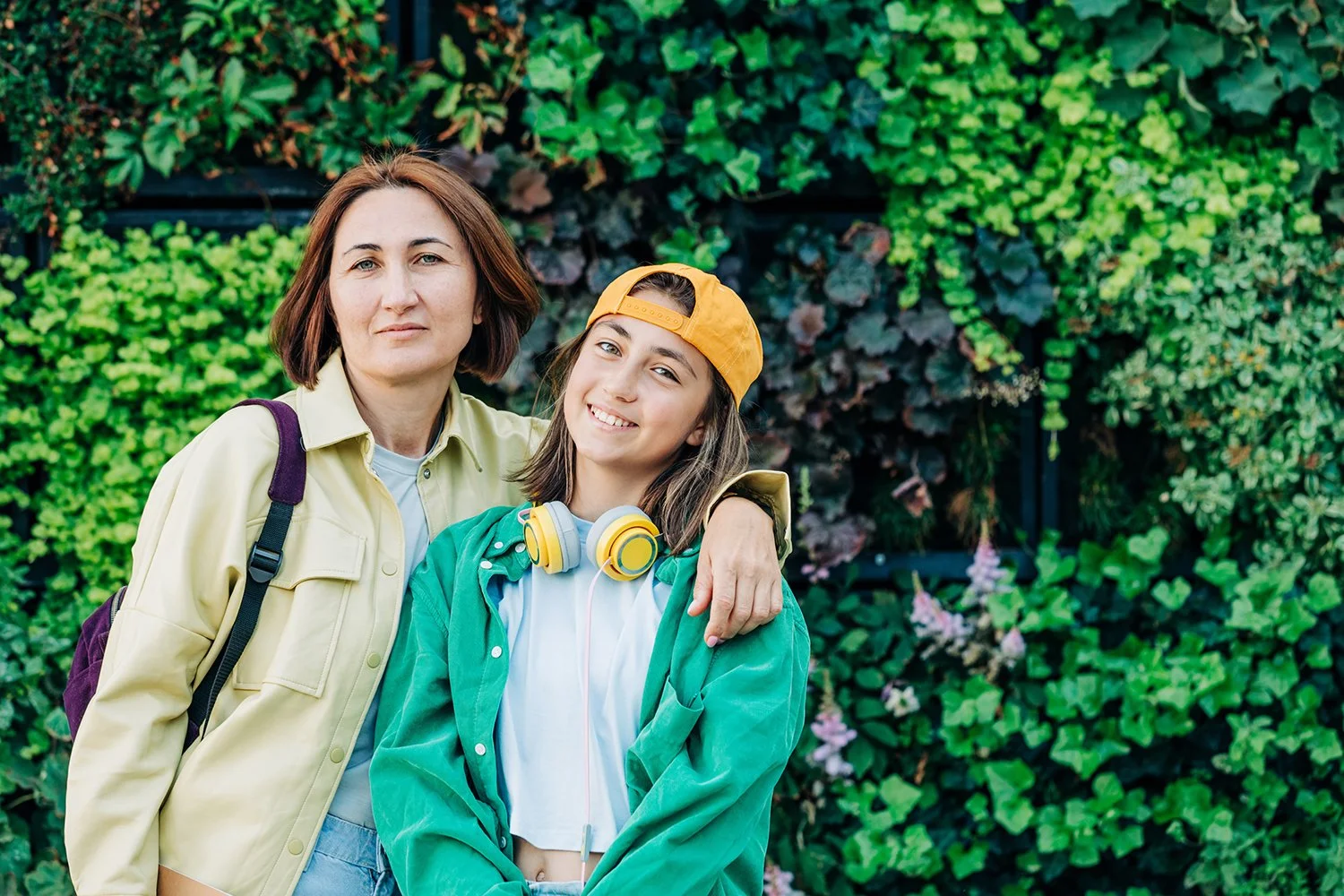 A woman with short brown hair and a young girl with long brown hair, both smiling, outdoors in front of green leafy plants. The woman is wearing a light yellow jacket and carrying a backpack. The girl is wearing a yellow cap, headphone, and a green jacket.
