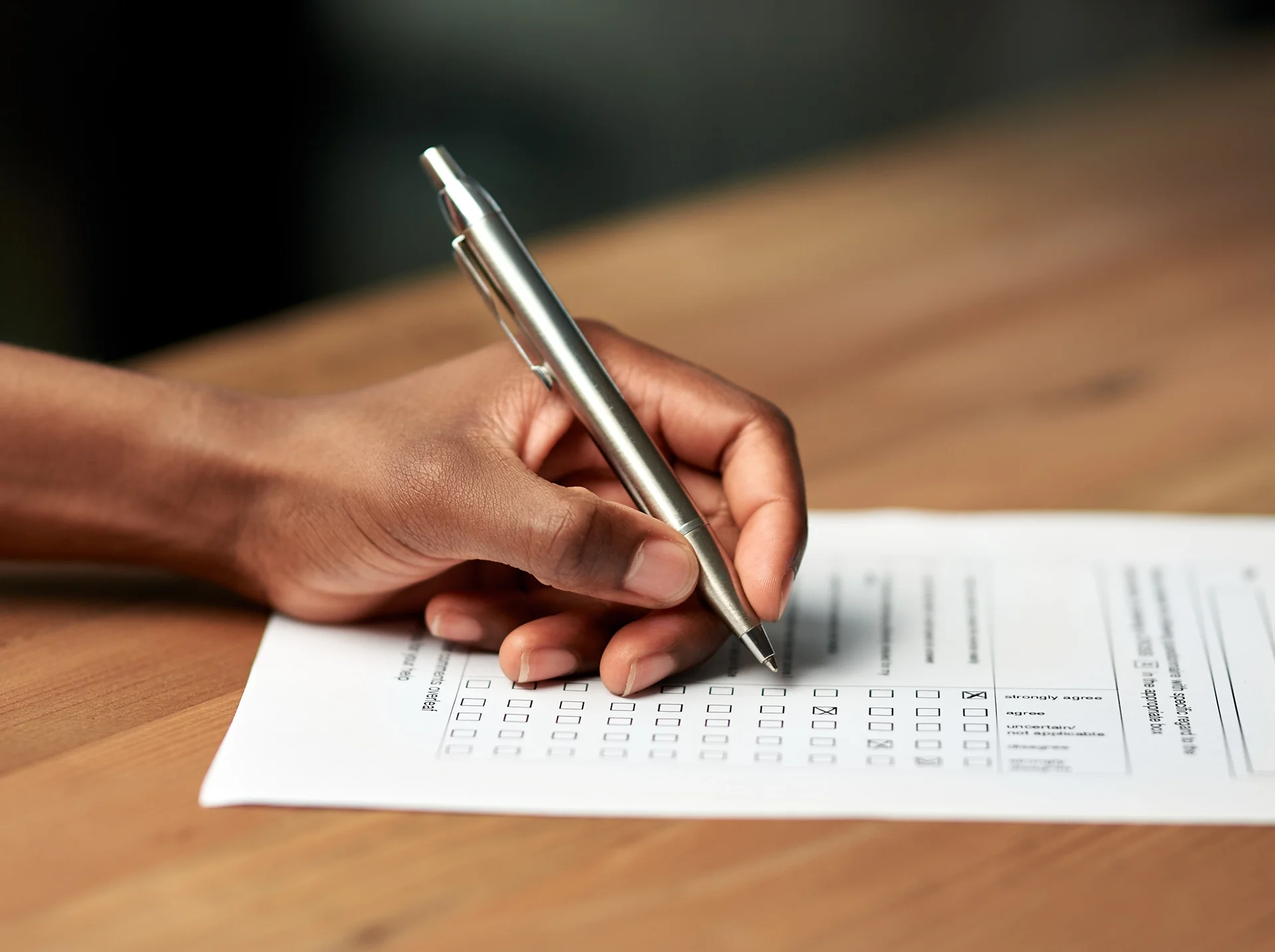 Close-up of a person's hand filling out a form with a silver pen on a wooden desk.