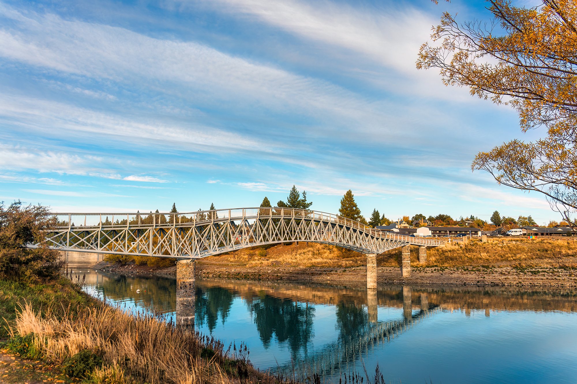 A white decorative pedestrian bridge crosses over a calm river, with a residential area and trees in the background, under a partly cloudy sky.