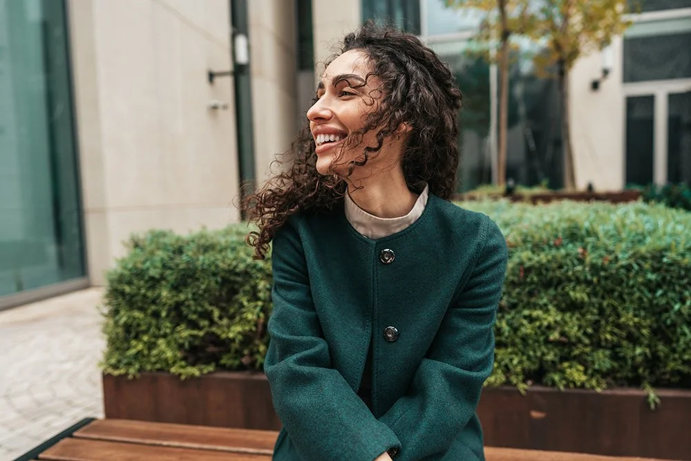 A woman with curly hair smiling and sitting outdoors on a park bench, wearing a dark green coat.