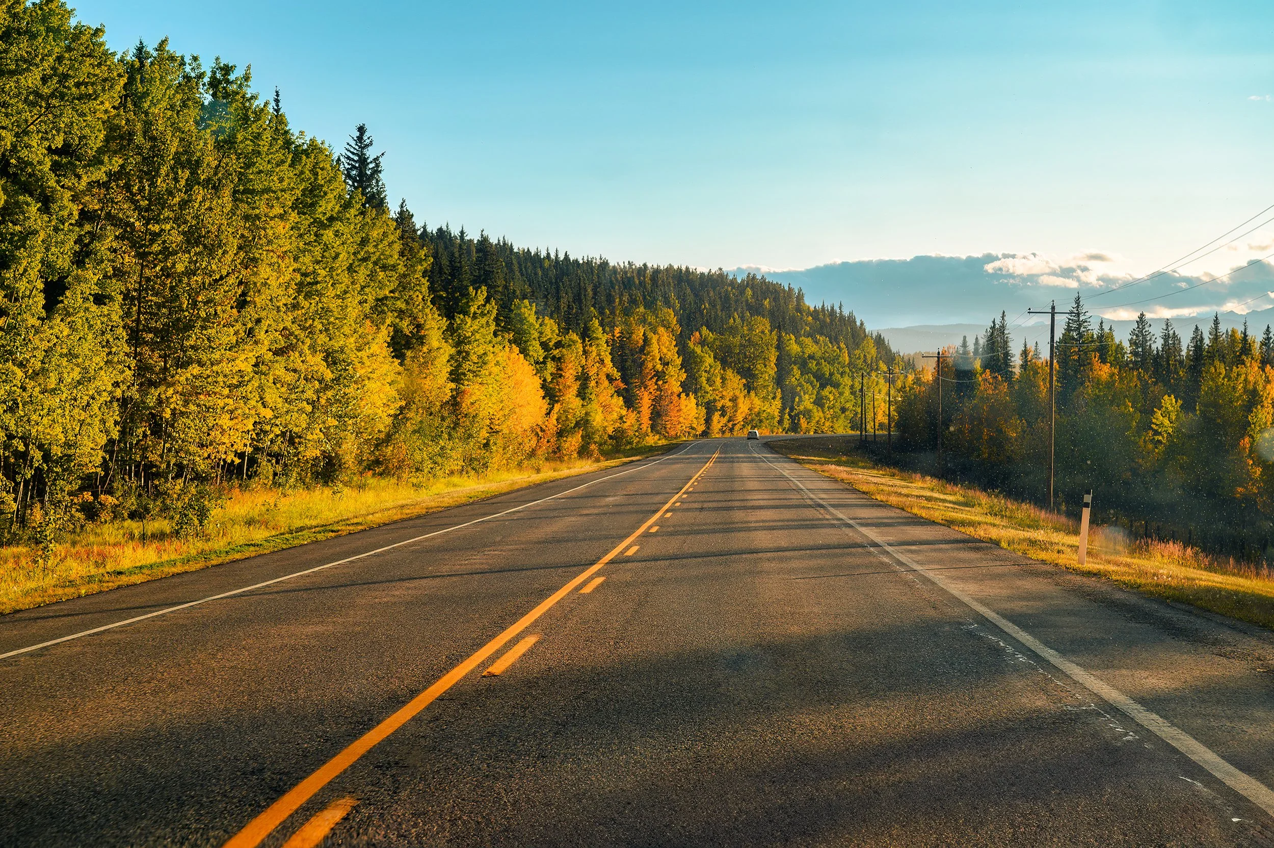 A two-lane highway running through a scenic landscape with trees in fall colors and a clear blue sky.