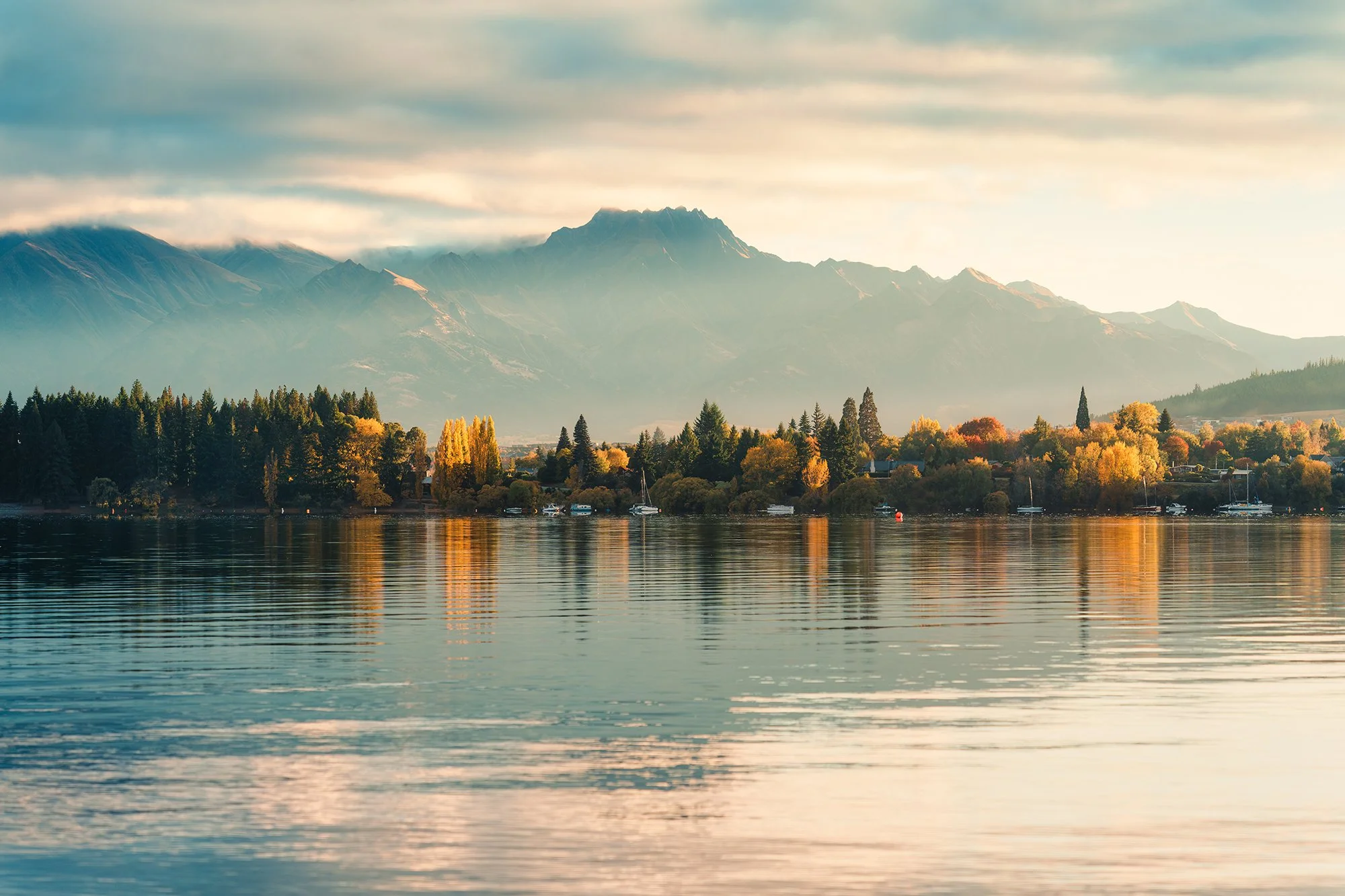A peaceful lakeside scene with calm water reflecting colorful autumn trees, distant mountains partially covered with clouds, and a partly cloudy sky.