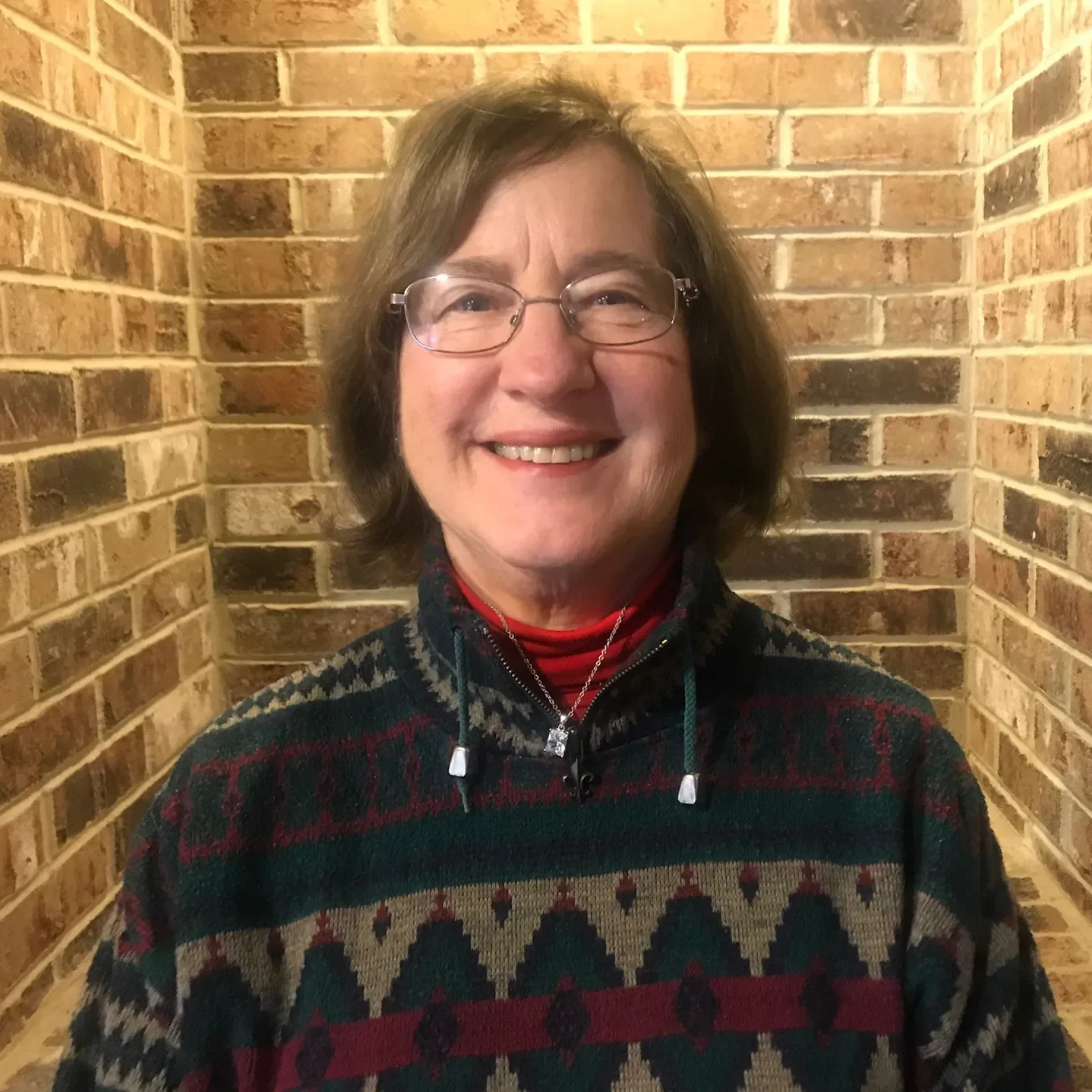 Smiling woman with glasses wearing a patterned sweater and a necklace, standing in front of a brick wall.