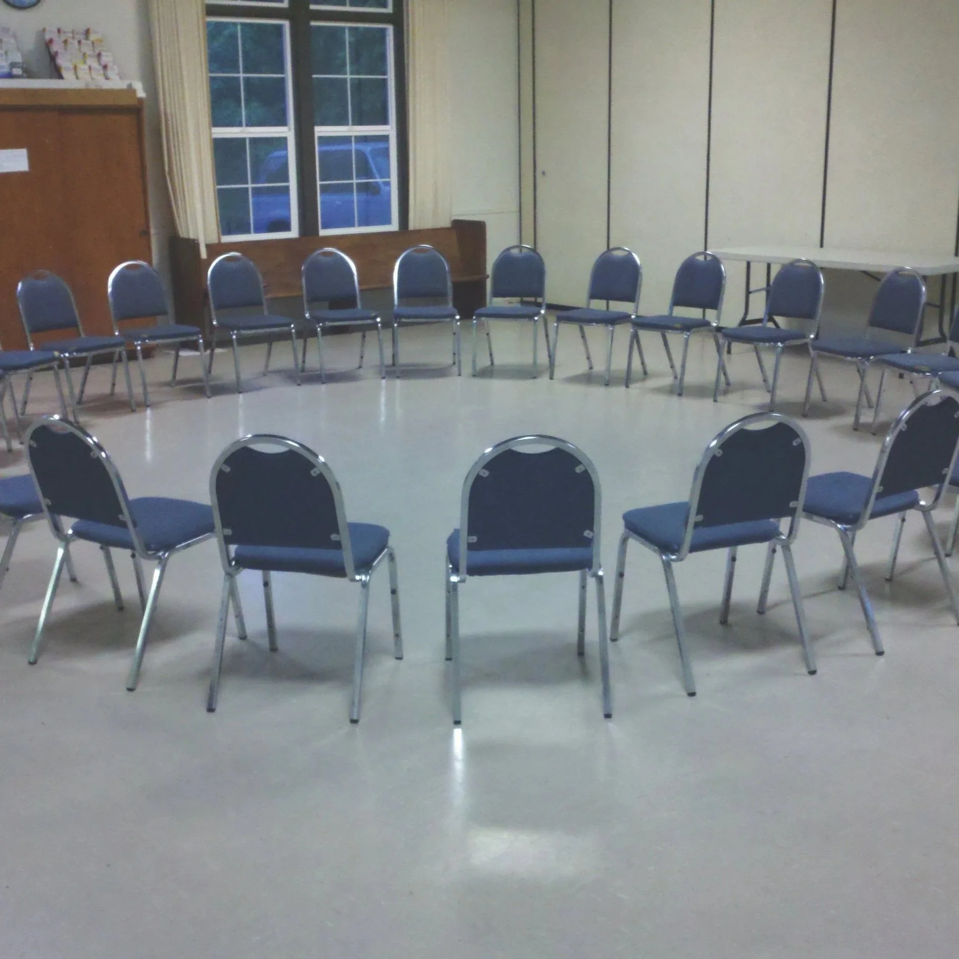 A circle of 14 blue padded chairs with metal frames in a room with beige walls, a window with white curtains, and a window bench next to the window.