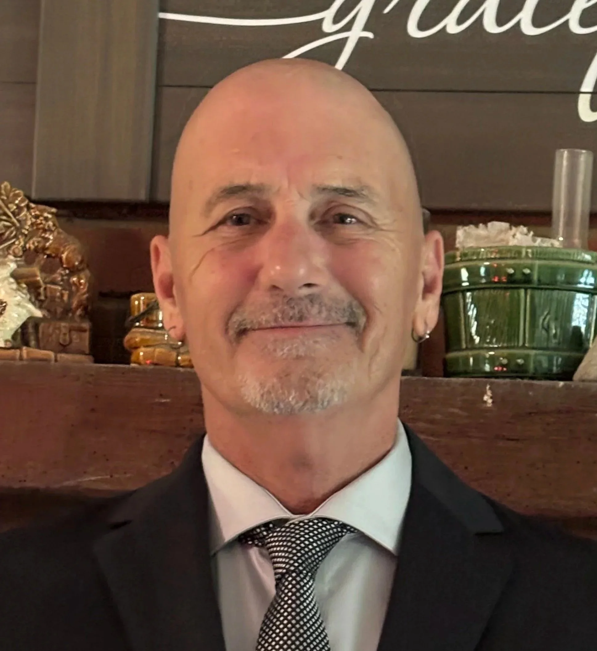 A bald man with a short gray beard and mustache, wearing a suit and tie, smiling at the camera, with decorative items and a green container in the background.