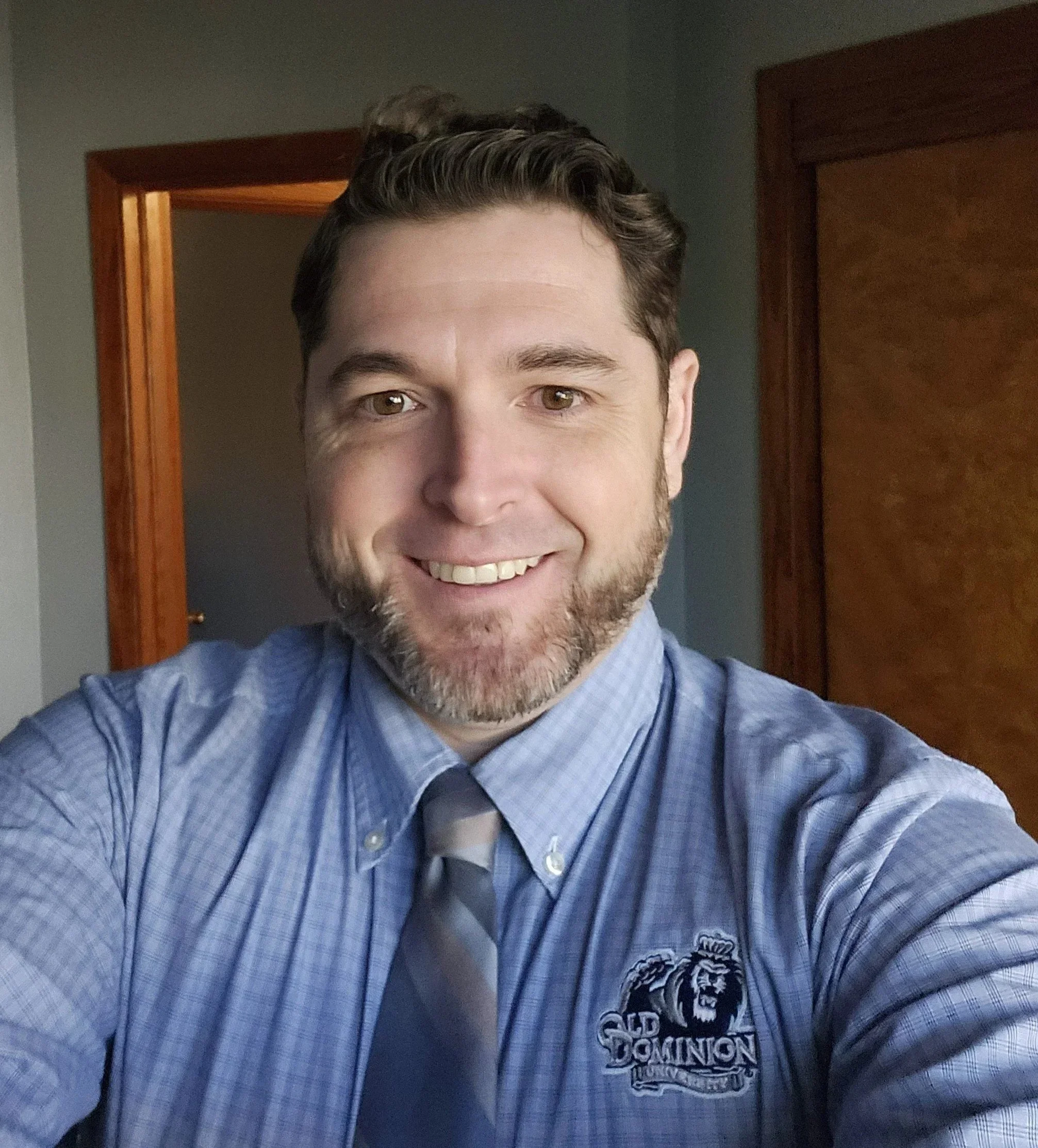 A man taking a selfie indoors, wearing a blue checkered dress shirt with an 'Old Dominion' logo and a gray striped tie, with a wooden doorframe and door in the background.