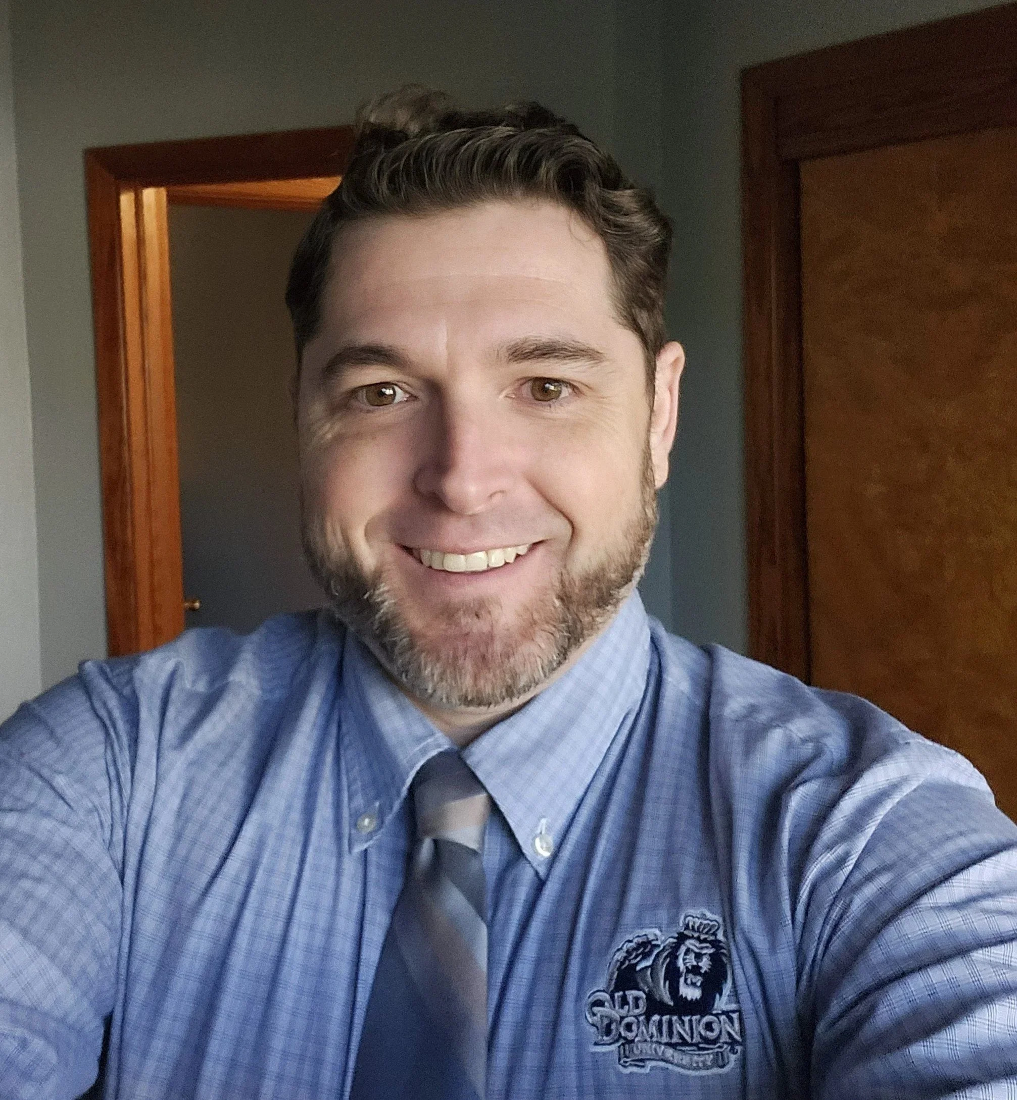 A man with brown hair, a beard, and light skin taking a selfie. He is smiling and wearing a blue dress shirt with a logo that says 'Old Dominion University' with a lion. He is also wearing a grey and white striped tie. The background shows a room with a wooden door and grey walls.