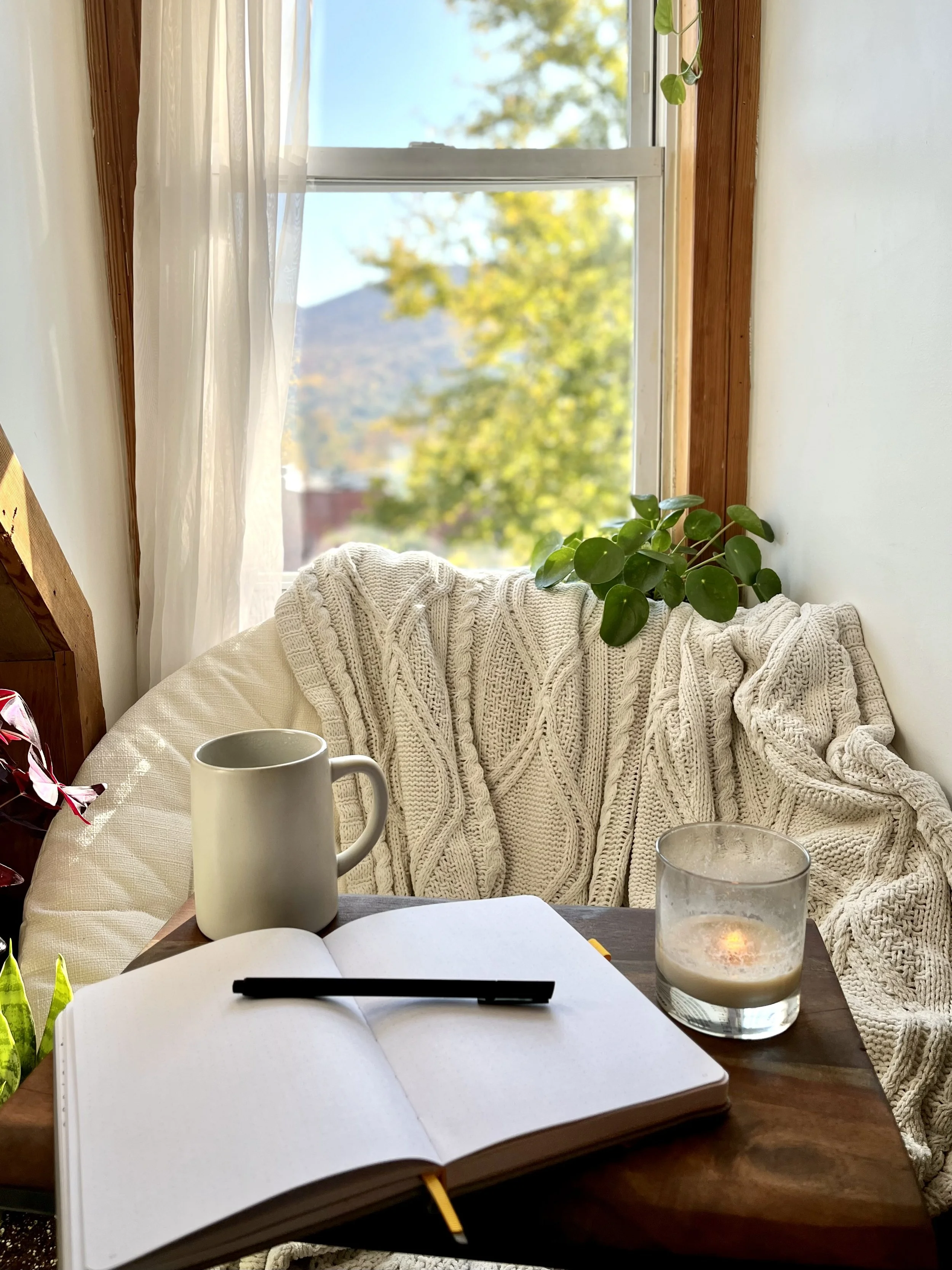 A cozy scene with an open notebook and a black pen on a wooden table, a white mug, a lit candle in a glass holder, a potted plant with green leaves on the windowsill, a cream-colored chunky knit blanket draped over a sofa, and a window showing an outdoor view with trees and mountains.