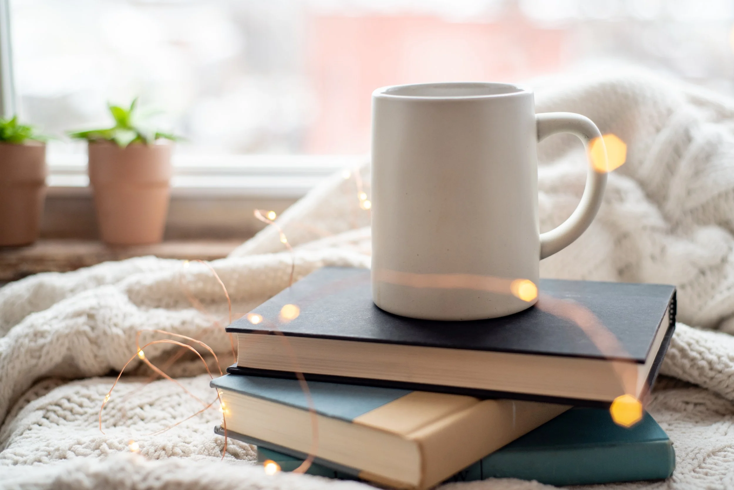 A white ceramic mug placed on top of a stack of books, with a window in the background. Image representing connection, community, and support through learning and wellness coaching.