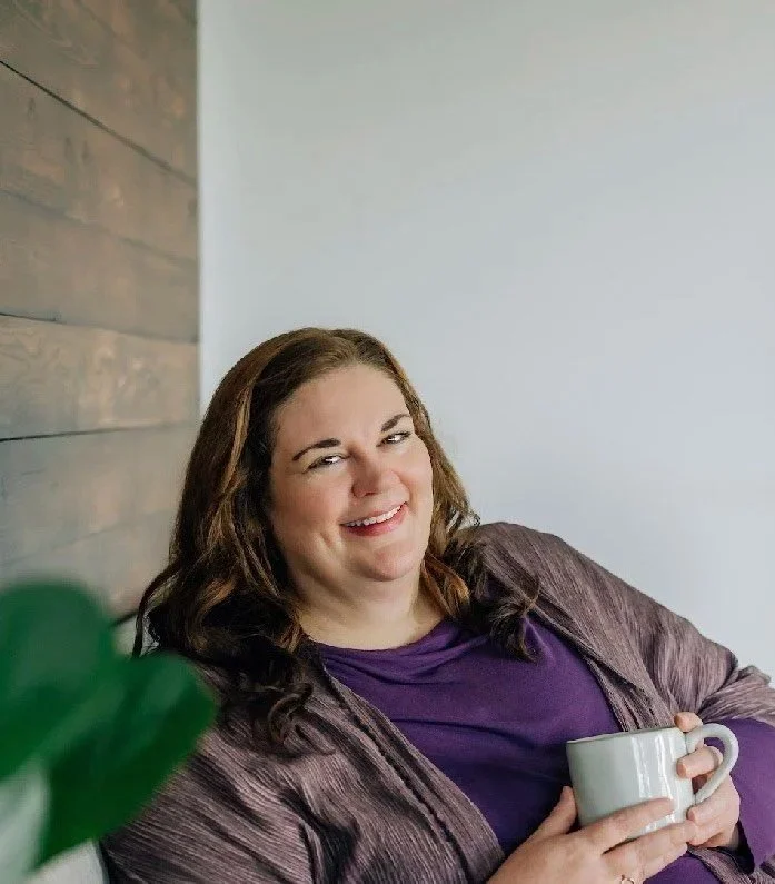 A woman with brown hair smiling and holding a white mug, sitting indoors near a wooden wall and a white wall, with some green foliage in the foreground.