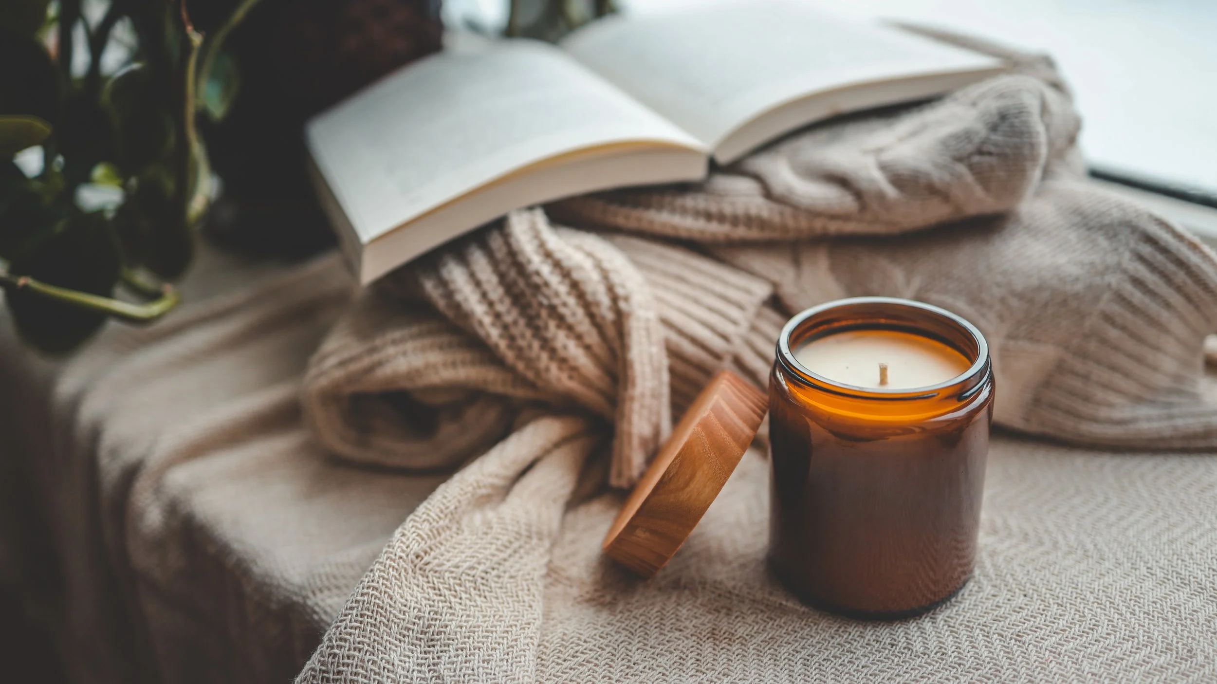 Open book resting on beige knitted blanket, next to a white candle in a brown glass jar, on a textured surface.