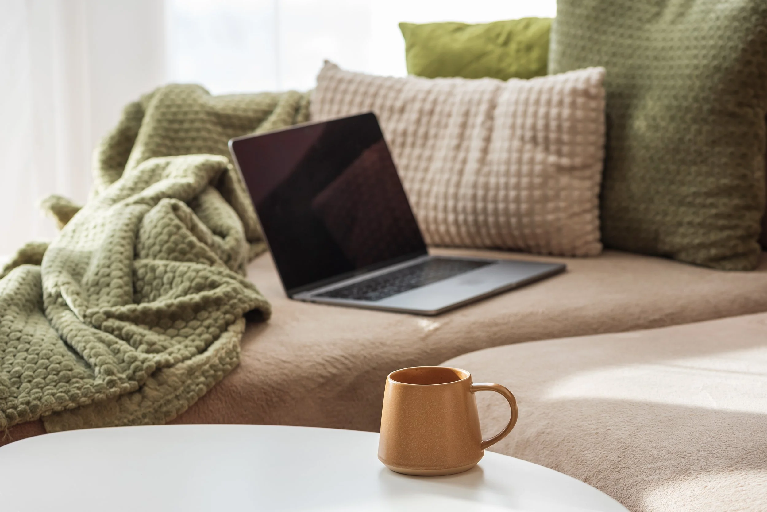 A beige sofa decorated with textured green and beige pillows, a green blanket, a laptop, a brown mug, and a white surface in the foreground. Sunlight filters through a sheer curtain in the background.